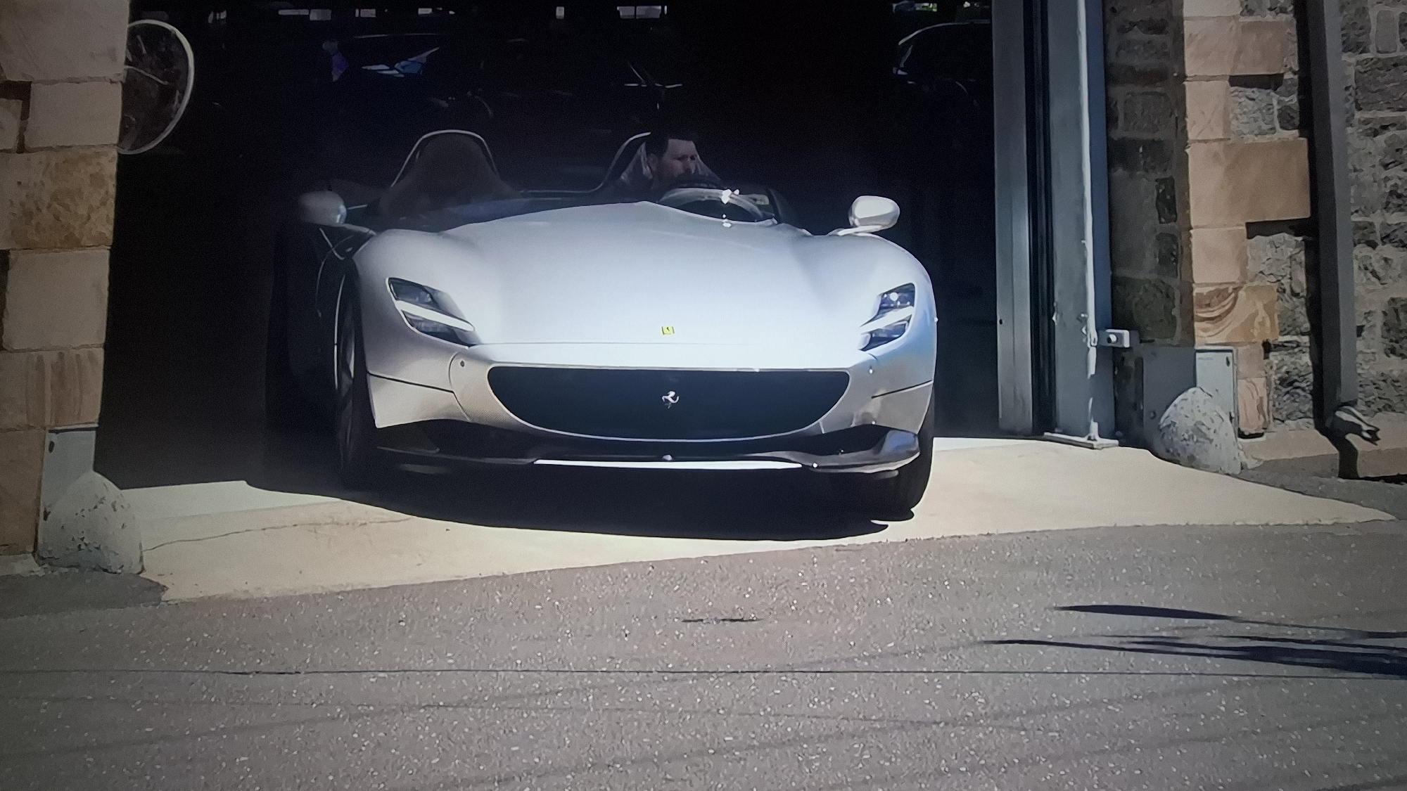 The image shows a silver Ferrari Monza SP1 parked in the open garage of a stone building. A man is seated in the driver's seat. Additional vehicles are visible in the background within the garage. The location is in the city of Unknown, Unknown.