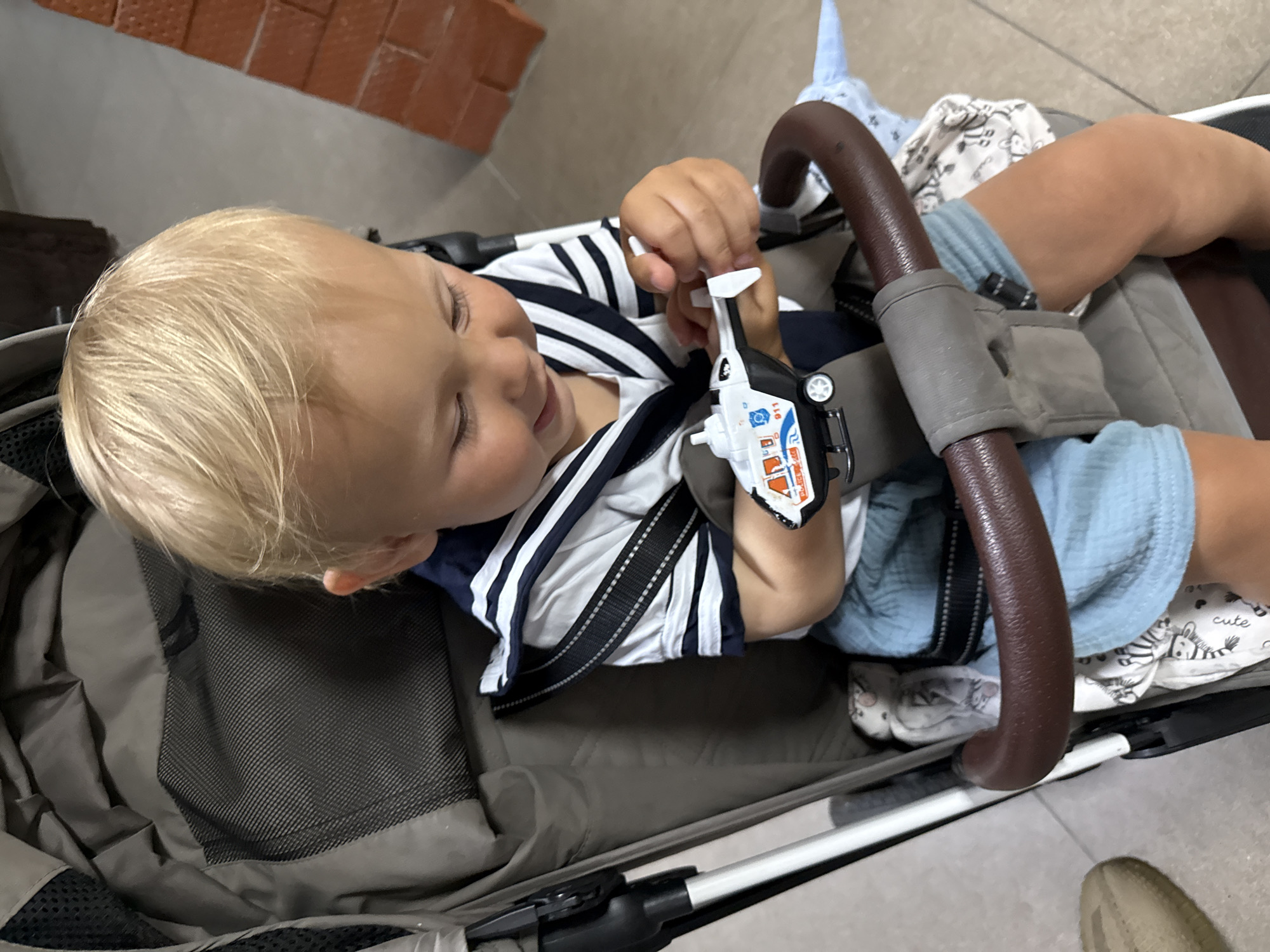 A young child with blond hair is lying in a stroller, holding a toy helicopter. The child is wearing a navy and white striped shirt and light blue shorts. The stroller has a brown handlebar and a gray fabric seat. A small, white toy helicopter with orange and blue details is in the child's hand. The background appears to be indoors, with a brick wall visible in the upper left corner. The floor is tiled. The child's eyes are closed, and they appear relaxed and content. There is a small amount of text on the toy helicopter, possibly "911". A patterned blanket with zebras and text reading "cute" is visible in the stroller. The image is brightly lit, suggesting it was taken during the day.