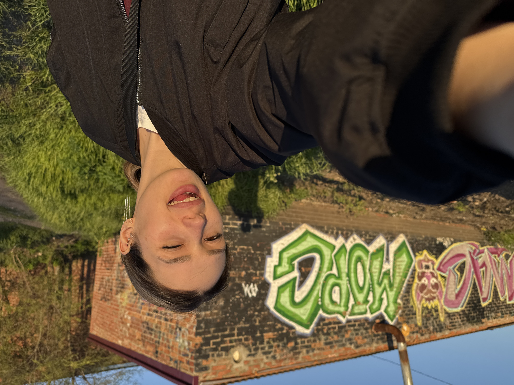 An outdoor, slightly tilted photograph captures a young person smiling joyfully, with eyes closed and teeth showing. They are wearing a black bomber jacket over a white top, and have long, delicate earrings on one ear. The warm, directional light suggests late afternoon or golden hour, creating a bright and pleasant atmosphere.
The background features a brick building, painted black at the top, covered in colorful graffiti. To the left, the graffiti is in green bubbly letters, spelling "MOW". To the right, in pink and purple hues, it spells "DOWN", with a distinctive reddish-brown and yellow cartoonish figure between the two words, suggesting the full phrase is "MOW DOWN". Below the graffiti, the brickwork is visible, hinting at a weathered structure. Beyond the building and to the person's left, there's green grass and some sparse, bare-branched trees or bushes, indicating an outdoor, possibly urban or semi-urban setting with some unkempt natural elements. The sky is clear and blue.