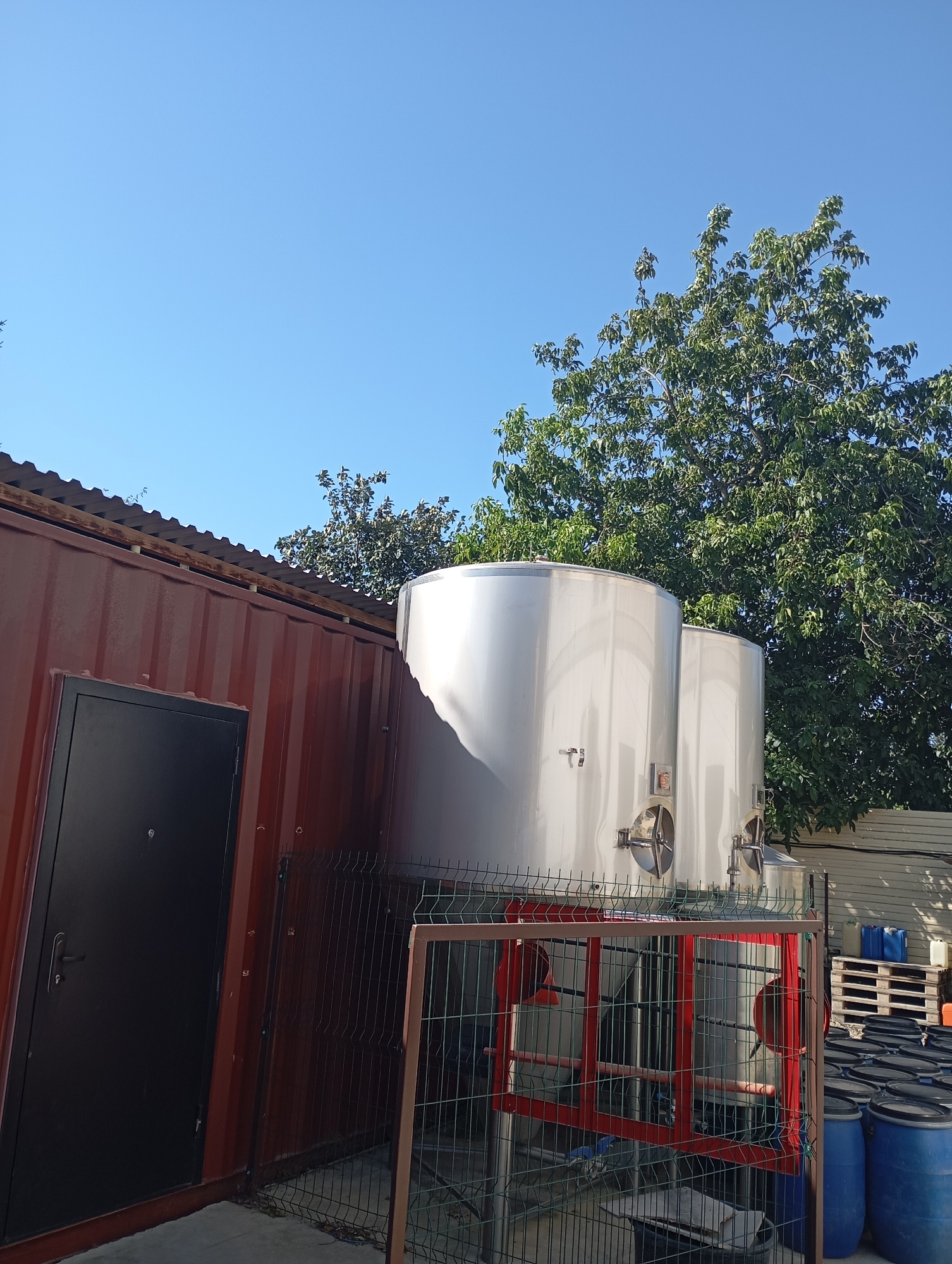 In Lazurny, Russia, an outdoor scene depicts industrial equipment under a clear blue sky. The foreground features a weathered, dark-colored metal shipping container with a black door. Adjacent to the container is a fenced area enclosing two large, stainless steel tanks, supported by a red metal frame. These tanks appear to be part of a storage or processing system. Behind the tanks, a dense green tree with full foliage dominates the background, providing a contrast to the industrial elements. Further to the right, several blue and white plastic barrels are visible, some stacked on wooden pallets, suggesting a storage area for liquids. The overall environment appears to be a functional outdoor space for industrial or agricultural purposes.