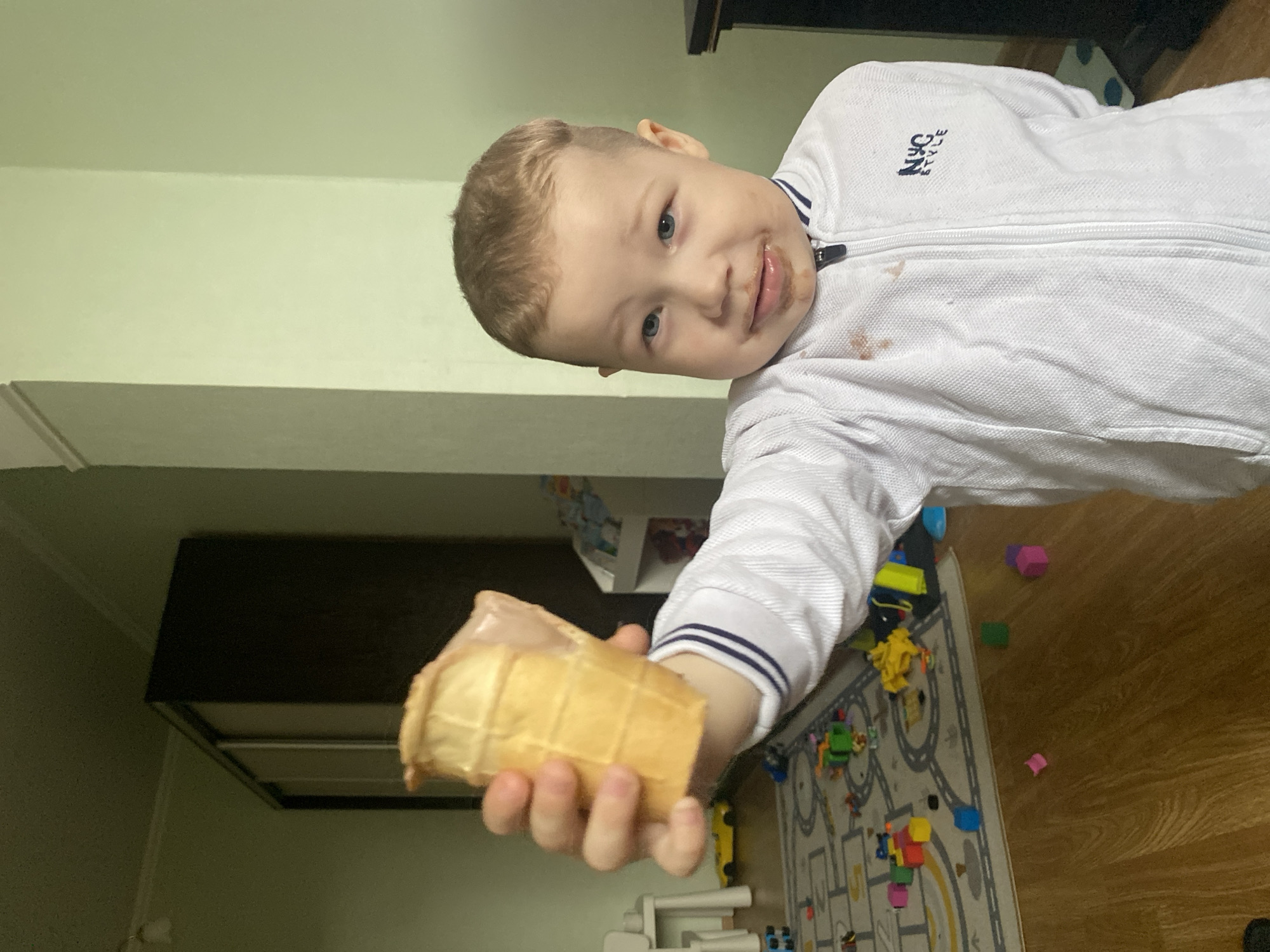 A fair-skinned boy with short blond hair is holding a partially eaten ice cream cone in his outstretched hand. The boy's face is smeared with chocolate, suggesting he was recently eating the ice cream. He is wearing a white zip-up sweatshirt with navy blue trim on the collar and cuffs, and the sweatshirt has some brown stains on it. The background includes a wall painted a light greenish-yellow color, with a brown dresser visible on the left side of the frame. A play mat with roads and toy cars is visible on the floor. There is no indication the photo was taken in Sochi, Russia.