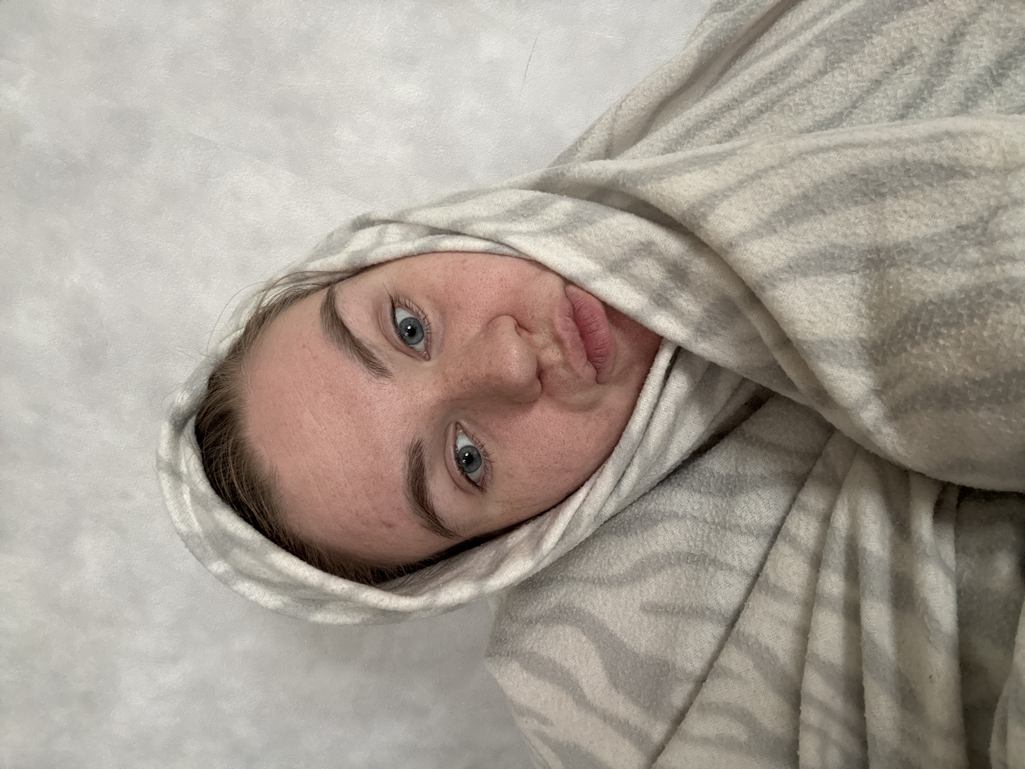 A fair-skinned woman with blue eyes and light-brown hair is depicted wrapped in a gray and white patterned blanket. The blanket covers her head, acting as a hood. Her face is turned to the side, and she is making a duck face. The background is a plain white surface. There is no location information available in this image.