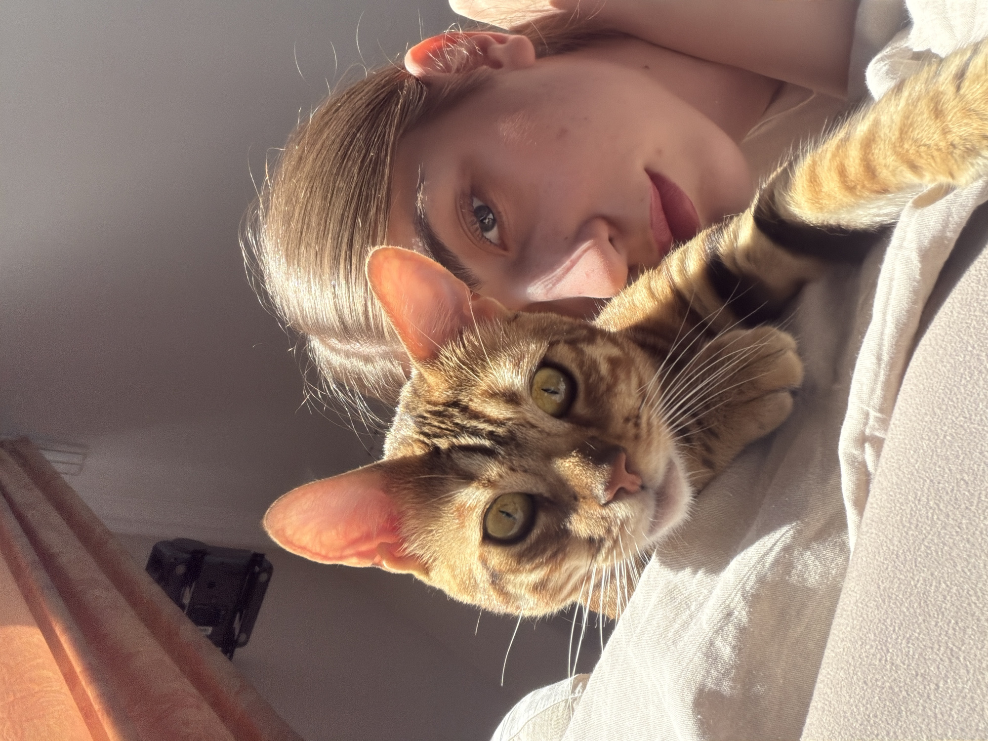 A young woman with light brown hair is lying on a bed next to a Bengal cat. The woman's face is partially visible, with her eye looking towards the camera. The cat is looking directly at the camera with green eyes. The sun is shining, creating strong shadows on the woman's face and the cat's fur. The background shows a curtain and a portion of a wall with a light fixture attached. There are no indications of location within the city of Krasnoyarsk, Russia.