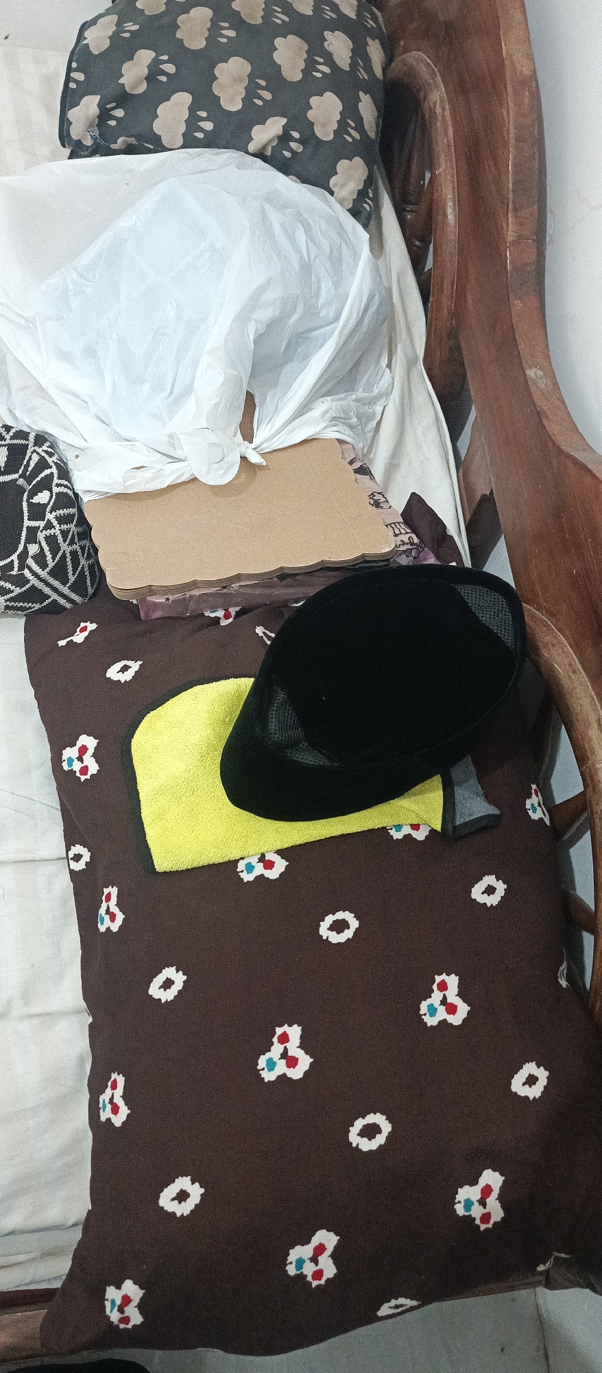 The image depicts a close-up view of a pillow with a brown fabric and a pattern of white, red, and blue floral shapes. A black cap is placed on top of the pillow, partially covering a yellow towel. Behind the pillow, there are stacks of items, including what appears to be a white plastic bag and some cardboard. A portion of a wooden chair is visible on the right side of the frame. The scene is likely set indoors, possibly in a bedroom or living area. The location in Arjasa, Indonesia, is not discernible from the visual elements within the image itself.
