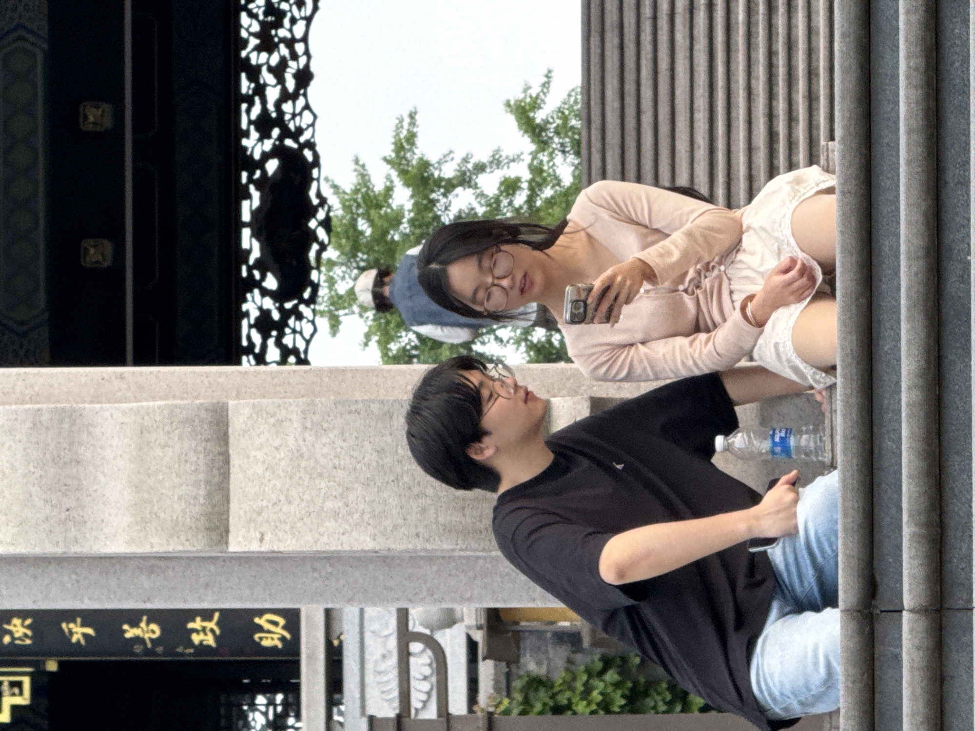 A young man and woman are sitting on a concrete structure in an outdoor setting, likely Liangjiang, China based on the traditional East Asian architecture visible. The woman, wearing glasses and a light pink top and white shorts, is looking at her phone. The man, wearing glasses and a black t-shirt with light blue jeans, is looking up at her. A water bottle is visible near the man. The architectural elements visible include grey concrete columns, steps and decorative latticework. The setting suggests a park or plaza.
