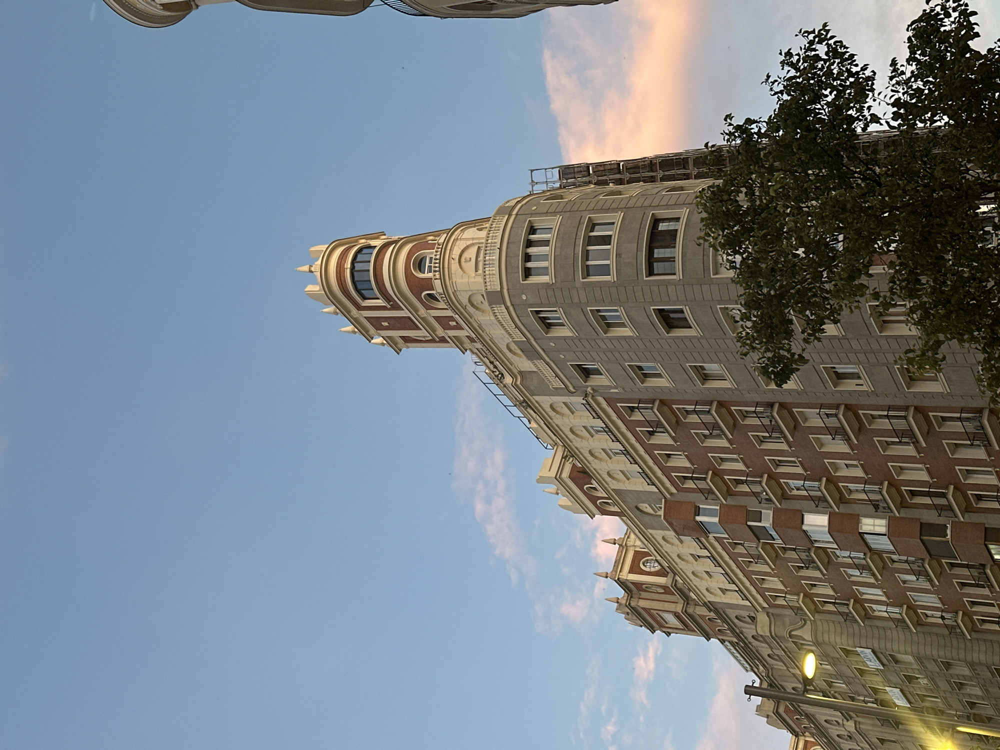 Here is a description of the image:
The photograph captures a low-angle view of a historic building in Madrid, Spain, ascending towards a light blue and pink-tinged sky. The building features a distinctive architectural style, with curved facades, intricate detailing, and a blend of colors, including grey, beige, and red brick. Its upper section includes a prominent, rounded corner with a decorated turret. A tree with green foliage partially obscures the upper right portion of the building. A street lamp is visible towards the lower right corner. Construction scaffolding is visible at the top part of the building. The sky shows signs of either sunrise or sunset, with the blue gradually fading into soft pink hues.