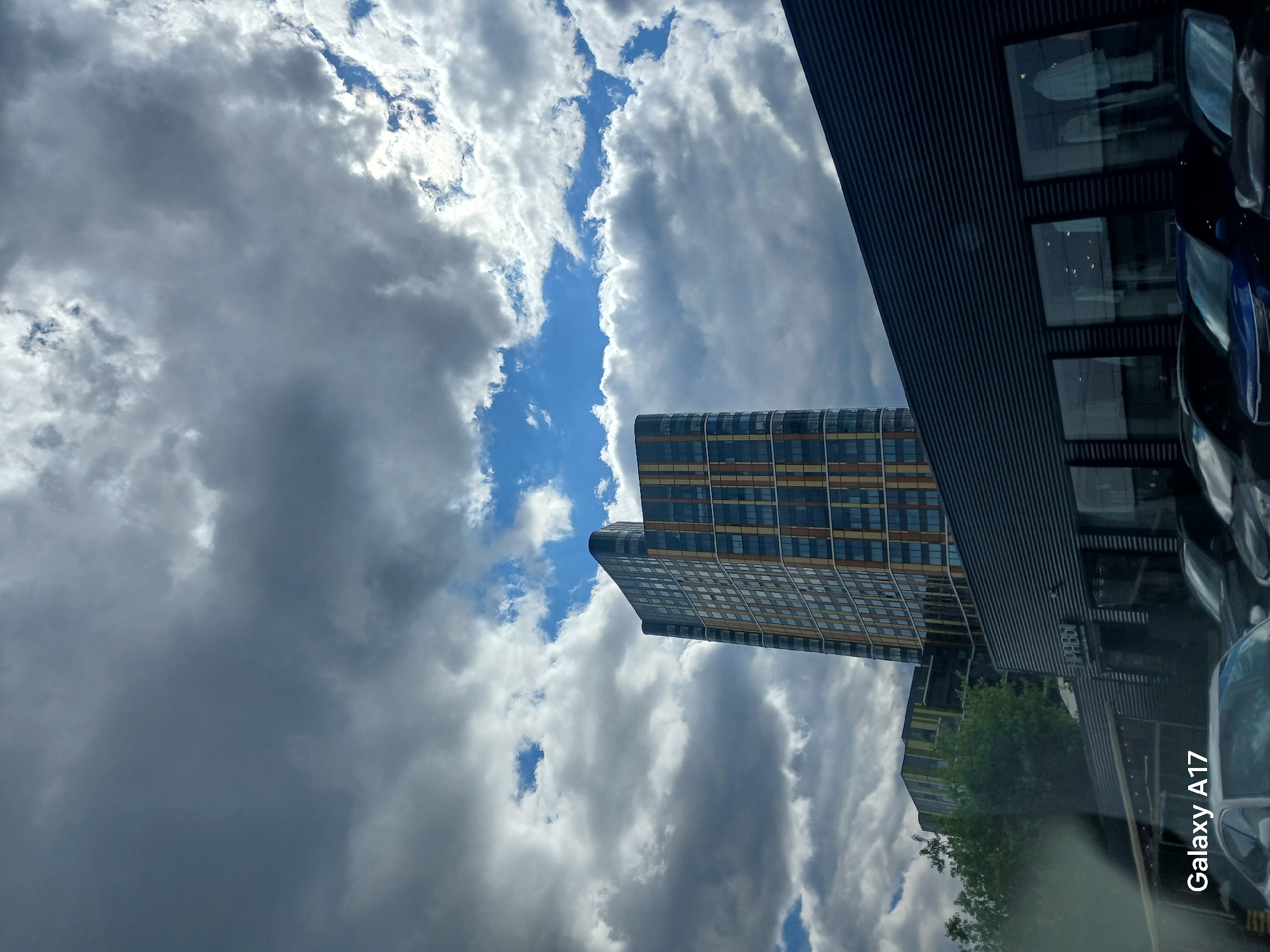The image shows a view looking up at a partially cloudy sky with buildings in the foreground, taken in Yekaterinburg, Russia. One notable building is a modern high-rise with a grid-like pattern of windows and gold accents. Adjacent to it is a lower, darker building with a ribbed exterior and windows reflecting the sky. The presence of cars suggests a street scene. The weather is overcast with patches of blue sky visible. The lower right corner shows reflection of car dashboard that suggests this view was taken from inside a vehicle. The words "Galaxy A17" are present in the bottom right corner.