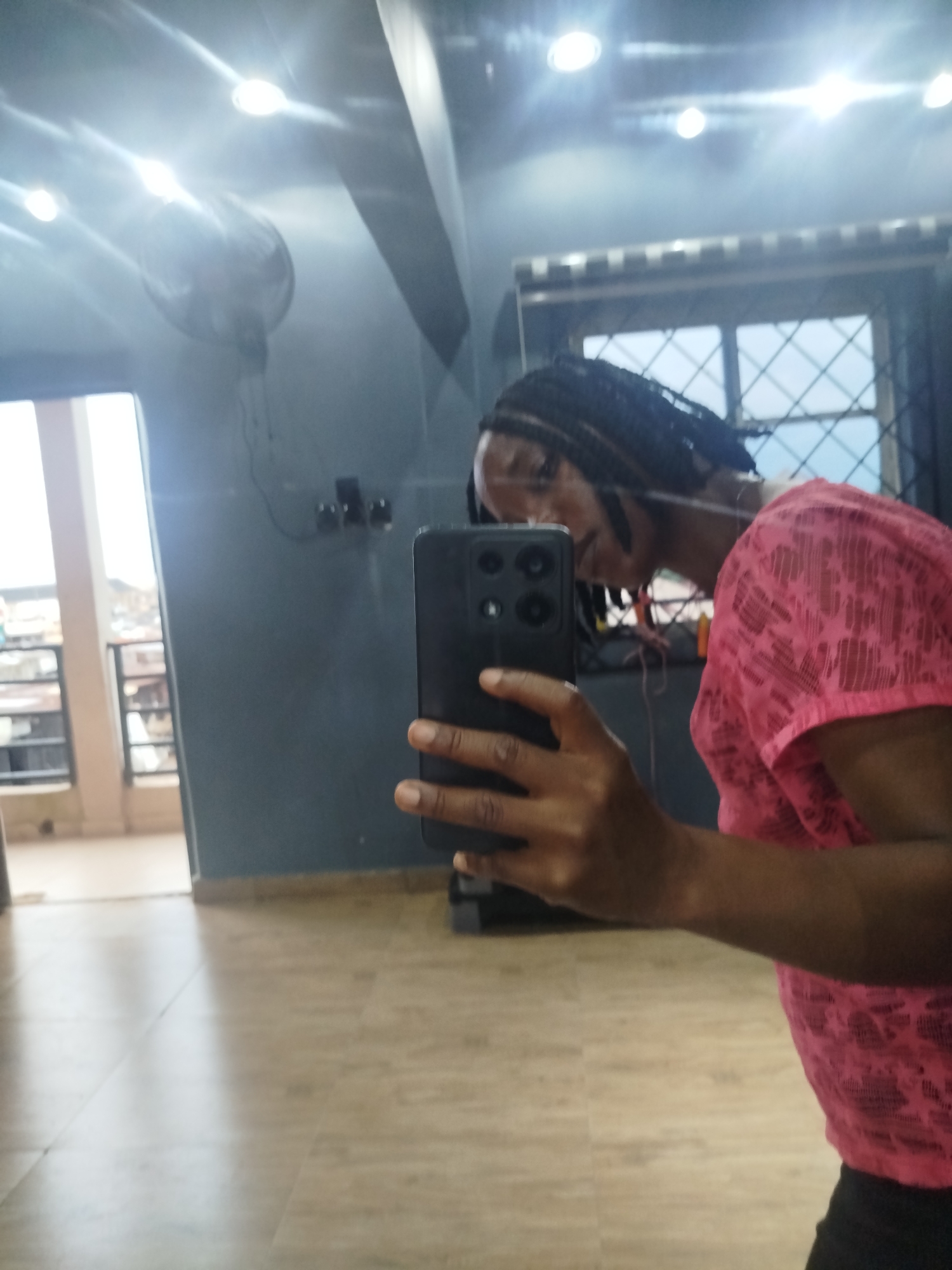 A medium shot captures a person taking a mirror selfie in what appears to be an indoor space with a reflective floor and a dark blue wall. The person, positioned in the right half of the frame, is wearing a pink, patterned, short-sleeved shirt and dark pants. Their dark hair is styled in braids that fall over their shoulder. They are holding a black smartphone in their right hand, angled to capture their reflection. The background shows a window with a crisscross pattern, a ceiling fan, and what looks like a doorway leading outside to a balcony area with buildings visible in the distance. The lighting in the room is bright, with several overhead lights visible. This scene is located in Uselu, Nigeria.