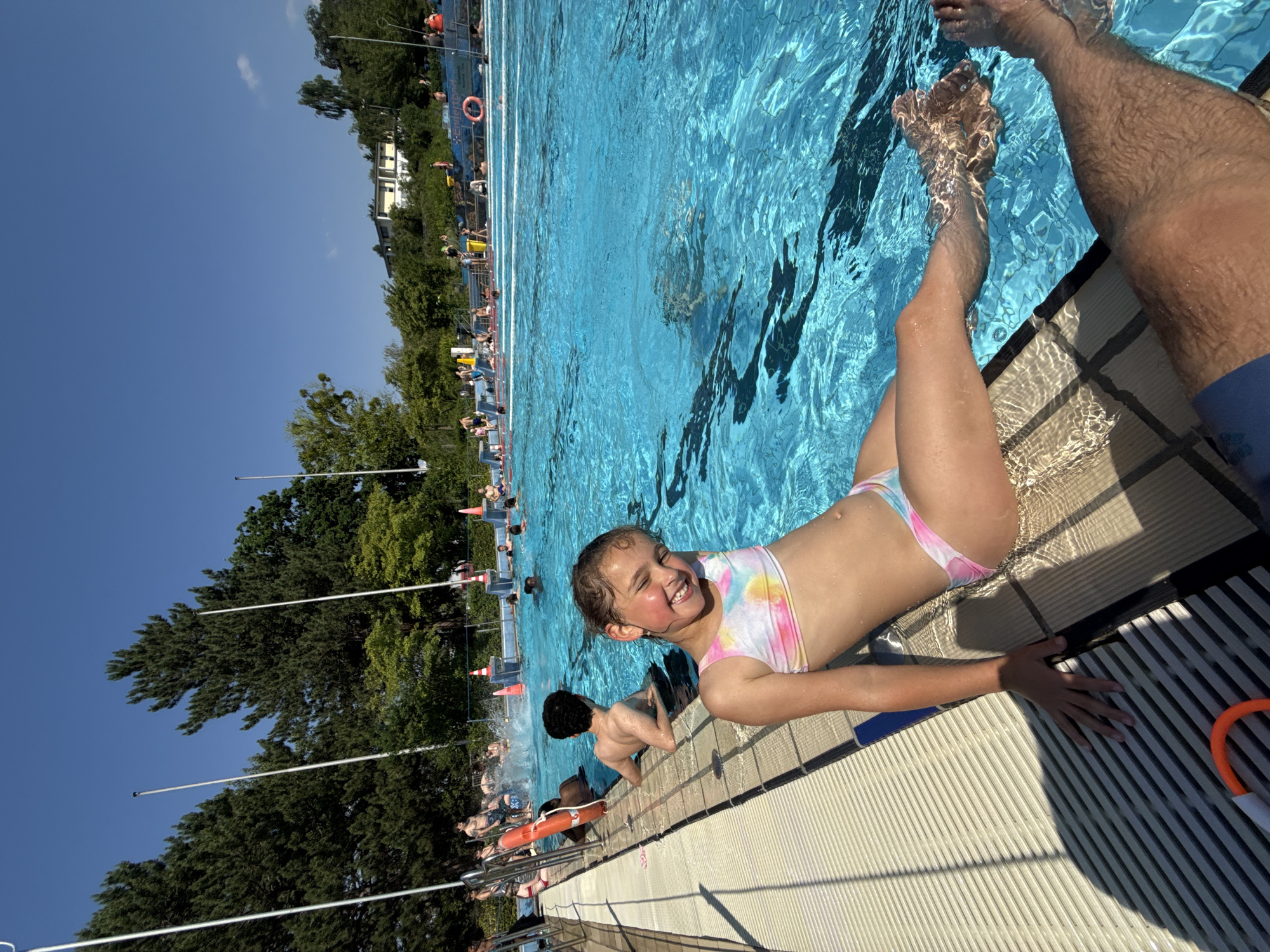 This is a photograph taken at an outdoor swimming pool in Bonn, Germany, on a sunny day. The image is oriented vertically, with the sky at the top left and the pool occupying the majority of the frame.
The primary subjects are two children. In the foreground, a young girl with a bright smile and a tie-dye bikini is lying on the edge of the pool, with her legs submerged in the water. Next to her, a boy with dark hair is sitting in the water, looking away from the camera. The water in the pool is a vibrant blue, with visible ripples and reflections.
In the background, other people are visible enjoying the pool and sitting by the edge. There are trees and buildings surrounding the pool, indicating an urban or suburban setting. Some pool equipment, such as lifebuoys and lane markers, are also visible. The time of day appears to be daytime, likely afternoon, given the bright sunlight and shadows. The mood of the image is cheerful and relaxed, with the girl's smile conveying happiness and enjoyment.
The image is a candid shot, capturing a moment of leisure and fun at the swimming pool. The lighting suggests it is a clear and warm day.