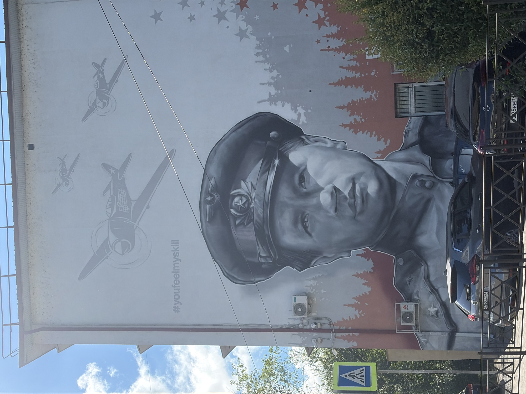 The image displays a large mural painted on the side of a multi-story building in Sochi, Russia. The mural features a grayscale portrait of a man in a military-style cap and uniform, with airplanes depicted above him. Text overlaid on the mural reads "#youfeelmyskill". The lower portion of the mural includes a silhouette of a forest and several stars. In front of the building, a street is visible with parked cars, a pedestrian crossing sign, and fencing. Trees and greenery are also present on the right side of the image. The sky is blue with scattered clouds.
