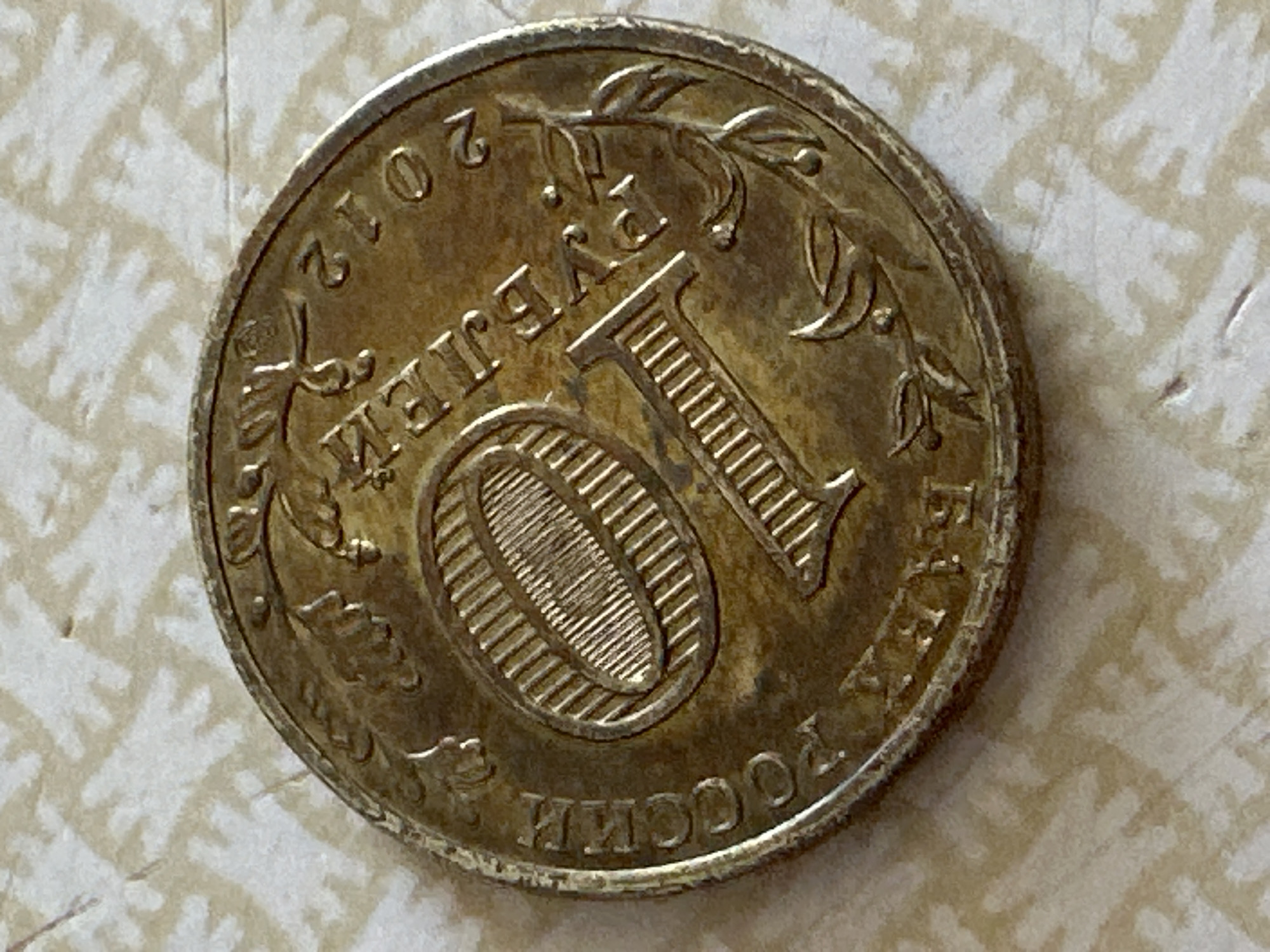 A close-up, top-down view shows a single, aged, golden-colored coin resting on a textured, light-colored surface with a repeating abstract pattern. The coin is circular with a raised rim and displays engraved details. Dominating the coin's surface is the large numeral "10," with intricate, parallel lines filling the oval shape of the "0." To the left of the numeral, curved lettering reads "РОССИИ." To the right of the "10," more curved lettering includes "РУБЛЕЙ" and the year "2012." Decorative elements, resembling branches with leaves and acorns, flank the central design. The coin appears to be a Russian 10 Ruble coin. The background surface is out of focus and has a slightly rough texture. There is no identifiable information regarding the city of Biysk, Russia, within the image.