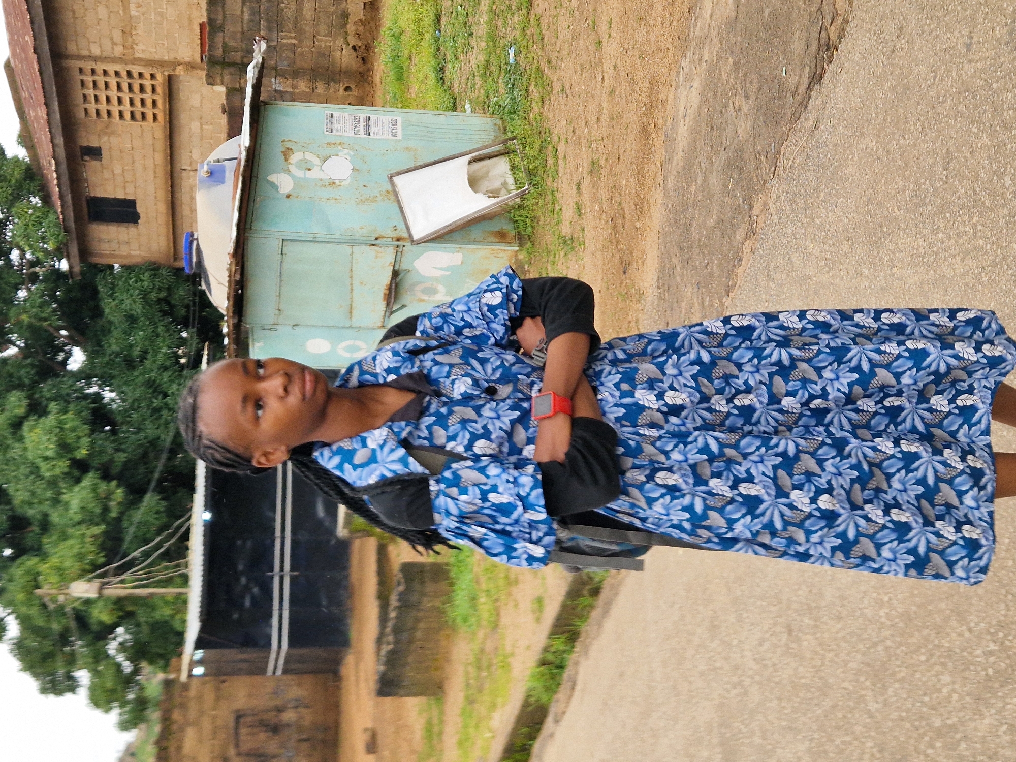 A young female, appearing to be of African descent, stands on a paved road in Kabong, Nigeria. She has dark skin and her hair is styled in braids. She is wearing a black long-sleeved shirt underneath a blue, white, and gray patterned dress with puffed sleeves. A gray backpack is visible on her shoulders. On her left wrist, she wears a red digital watch. Her arms are crossed.
In the background, there is a light blue metal kiosk with a light-colored water tank on top. To the left of the kiosk, a black structure with a corrugated metal roof is visible. Behind these structures, a two-story building constructed from light brown bricks stands. One window on the upper floor is dark, and another has a grid of ventilation holes. Trees with green foliage are present in the background, and the ground is covered with patches of dry grass and dirt. The overall lighting suggests an overcast day.