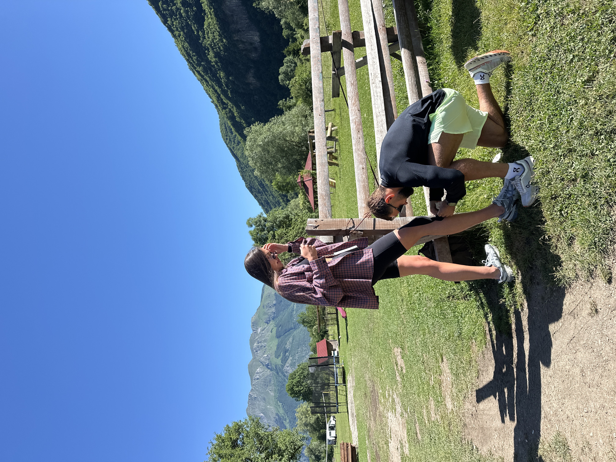 In Gusinje, Montenegro, on a bright, sunny day with a clear blue sky, a man kneels on green grass, adjusting the sock of a woman standing beside him. The woman, wearing black cycling shorts and a plaid shirt, looks down at her phone. A rustic wooden fence runs horizontally behind them. In the background, trees dot a grassy field, with mountains rising in the distance. A white camper van, a trampoline, and a small red-roofed building are visible further back, suggesting a campsite or recreational area. The overall scene depicts a moment of casual interaction outdoors in a scenic, mountainous environment.