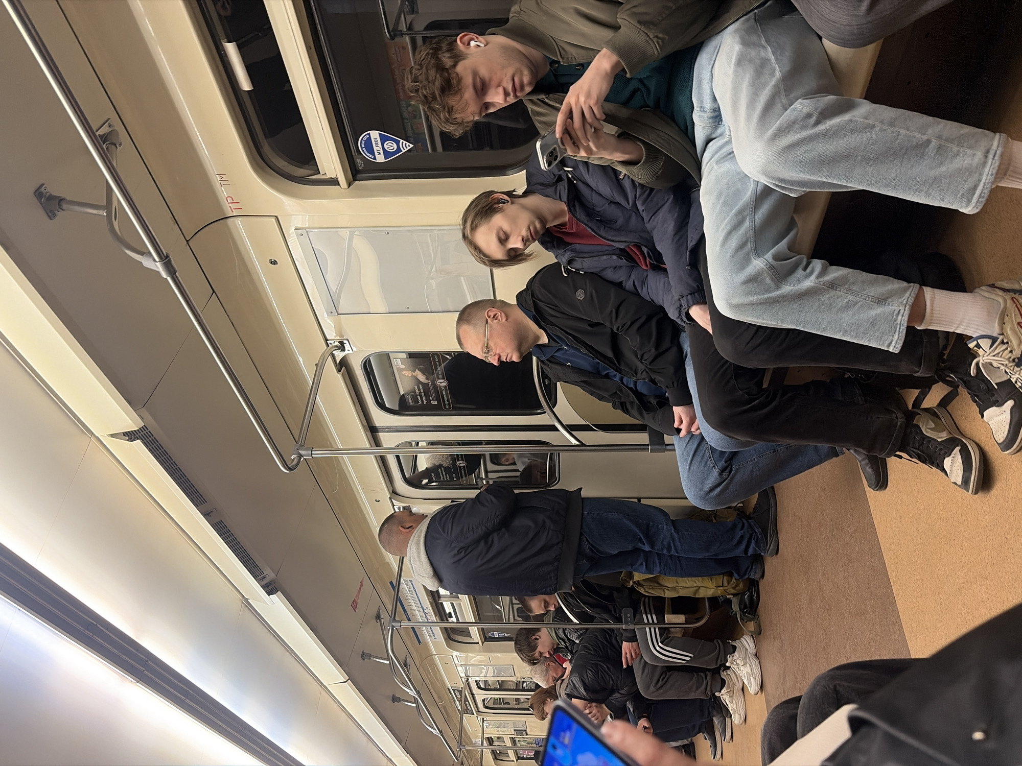 The image depicts the interior of a subway car in Saint Petersburg, Russia. Several passengers are visible, some seated and others standing. The seated passengers include three men in the foreground, with one using a smartphone. The interior features pale yellow walls, metal handrails, and various signs. The floor is brown. The carriage appears crowded. The passengers are dressed in casual attire.