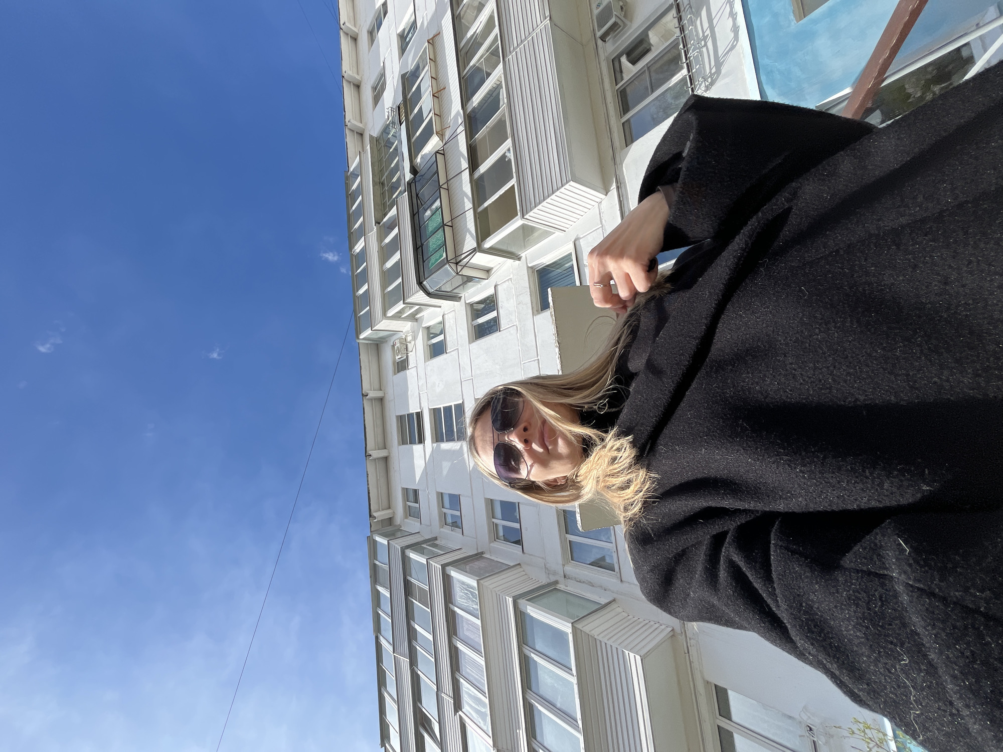 An outdoor, upward-angled portrait shot captures a person against a bright blue sky and a tall, light-colored building in Chaikovsky, Russia. The image is rotated clockwise, making the building appear to be rising diagonally from the lower left towards the upper right, with the sky filling most of the left side.
The subject is a person, likely female, positioned in the lower-right quadrant of the frame. They have blonde hair, partially obscured by the angle and sunglasses, and are wearing large, dark sunglasses that cover most of their eyes. A confident expression is conveyed. They are dressed in a dark, possibly black, heavy coat or jacket, which takes up a significant portion of the foreground. One hand, adorned with a ring on the ring finger, is visible near the person's neck, gently touching their hair or collar.
The background features a multi-story residential or apartment building with a white or off-white facade. Numerous rectangular windows, some with small balconies or bay windows, are visible along its structure. Several air conditioning units are mounted on the building's exterior. A hint of blue paint or paneling is visible on a section of the building towards the upper right.
The sky is a clear, vibrant blue with only a few wispy white clouds scattered across it, indicating sunny weather. A thin, dark wire or cable stretches across the sky in the upper-right portion of the image. The lighting suggests it is daytime, likely midday or early afternoon, given the brightness and minimal shadows. There is no visible text in the image.