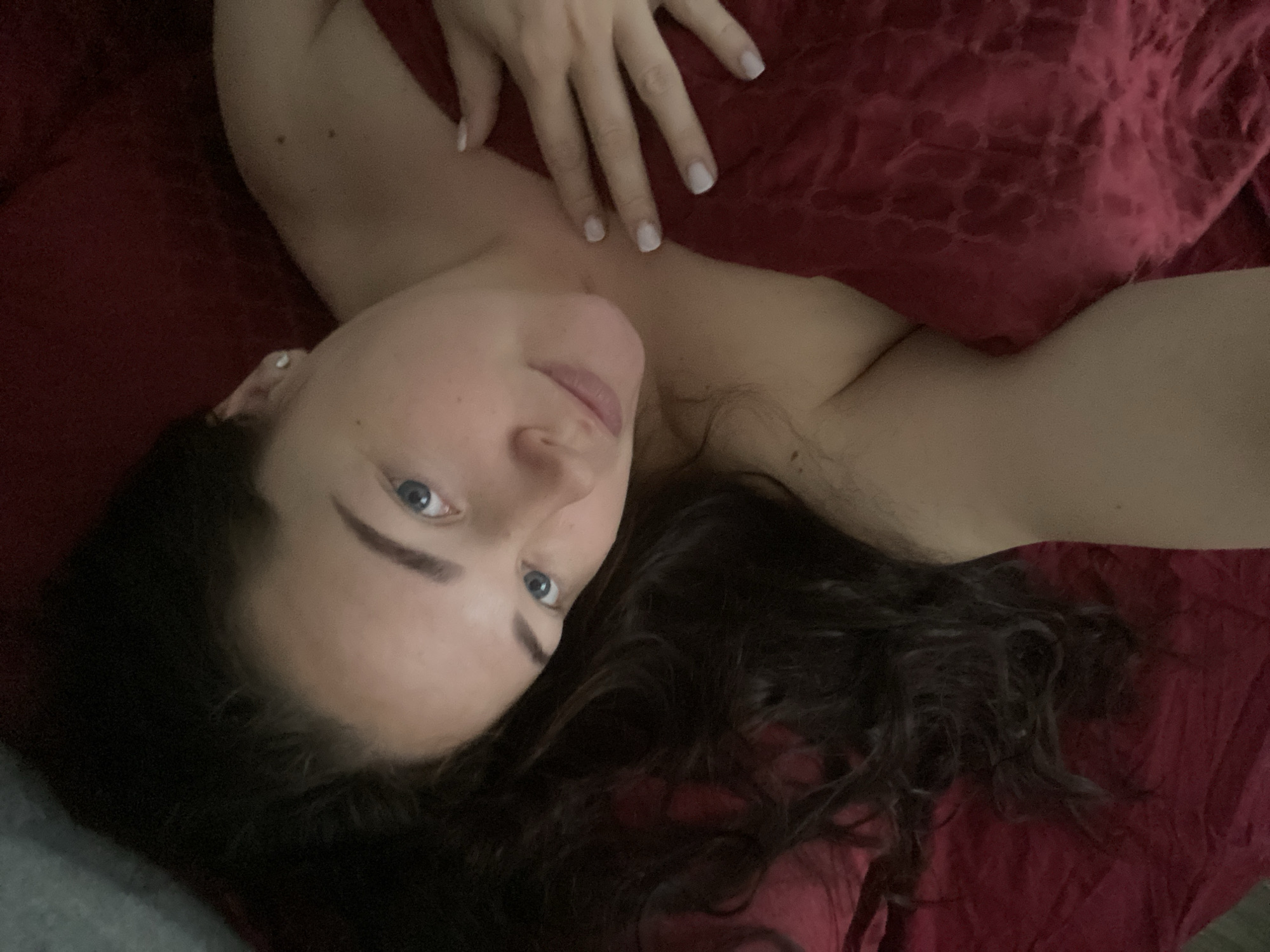 A woman with long, dark curly hair, fair skin, and blue eyes is lying on a red bedspread. Her right hand rests on her chest. The image suggests an indoor setting. No information indicates the location is Sochi, Russia.