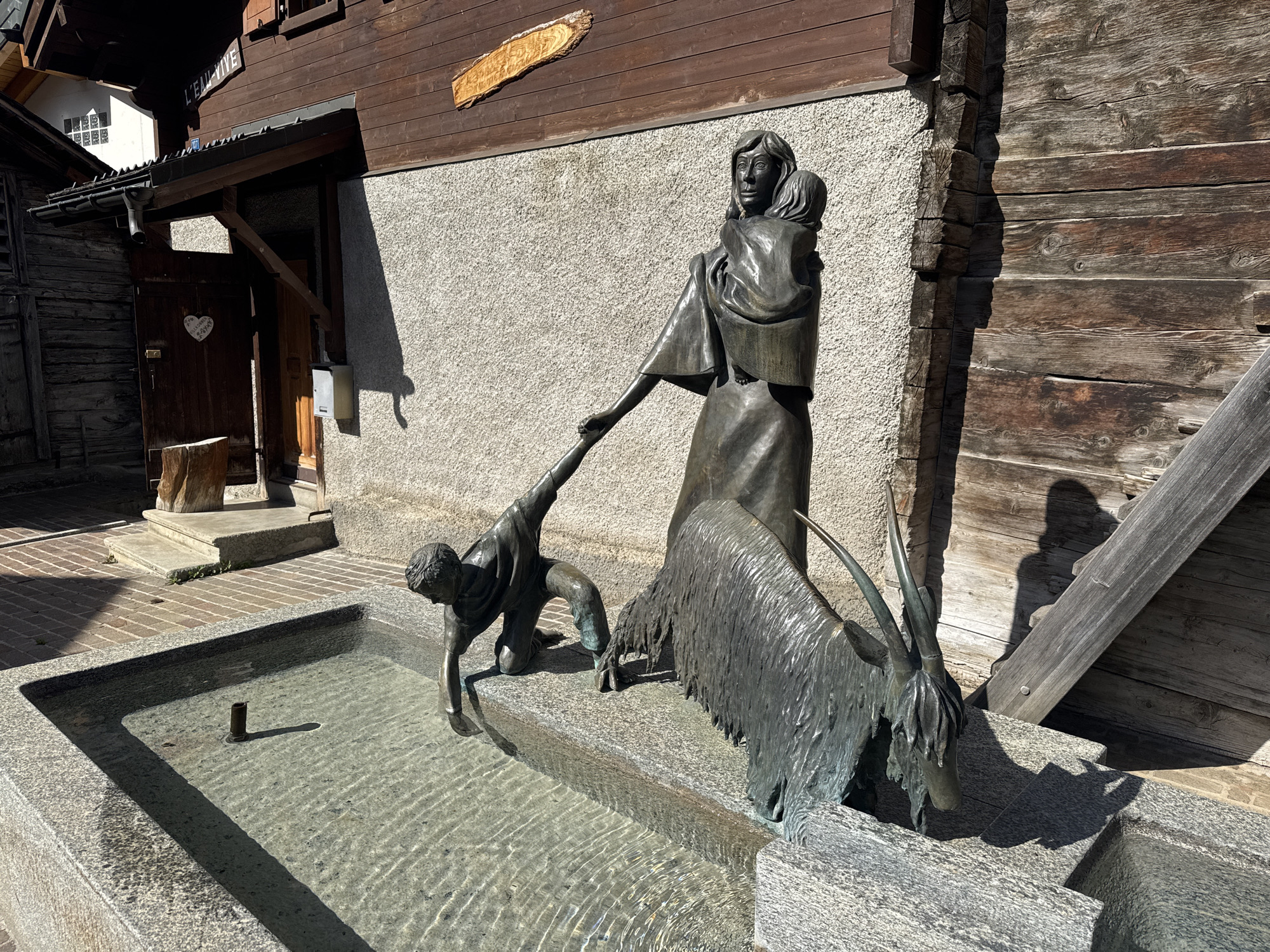 This is a bronze sculpture in a public fountain located in Leukerbad, Switzerland. The sculpture depicts a woman holding a child on her hip, reaching down to assist a kneeling child who is interacting with a goat. The goat is also depicted drinking from the fountain. The fountain is made of granite and is filled with clear water. The background consists of a textured stone wall and wooden buildings, typical of Swiss architecture. The scene is illuminated by bright sunlight, casting distinct shadows. The location is indicated by a sign reading "L'EAU VIVE" on the wooden building.