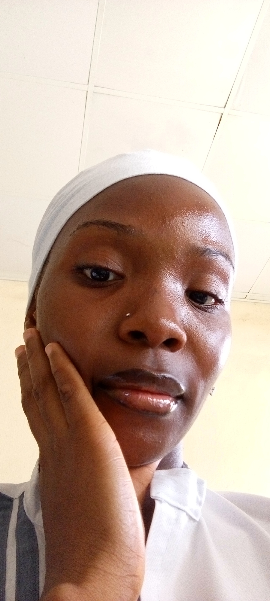 A close-up selfie depicts a person with dark skin and a white head covering. The person's left hand is raised to their cheek. They are wearing a collared shirt in white and grey stripes. The background is composed of an off-white ceiling, suggestive of an interior setting. The city and specific location are undetermined.