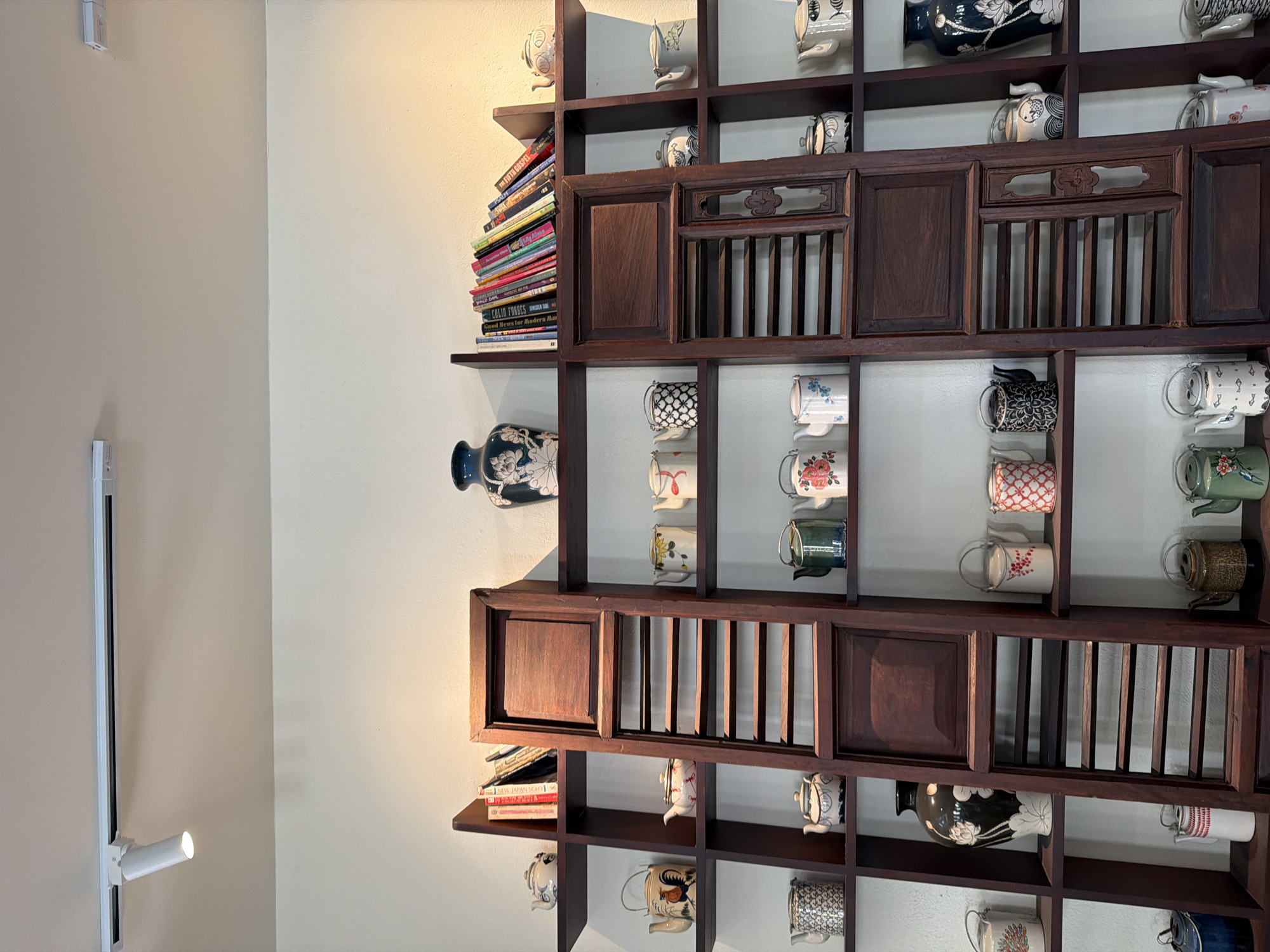 The image depicts a tall, dark wood shelving unit filled with various ceramic teapots and a few books. The shelves are organized in a grid-like structure. On the top shelf, a large blue and white vase with floral motifs is displayed. To its right, a collection of colorful books is arranged in a leaning fashion. Several teapots of different sizes, colors, and patterns are visible across multiple shelves, some with lids and handles. A decorative wooden panel, possibly part of an old window or door, is placed vertically in the center of the shelving unit. The wall behind the shelving unit is painted a light, neutral color. The lighting suggests an indoor environment, possibly a shop or a home display. Based on the context, this scene is located in Đà Nẵng, Vietnam, a coastal city known for its blend of traditional and modern aesthetics, where such antique decorative items are often found.