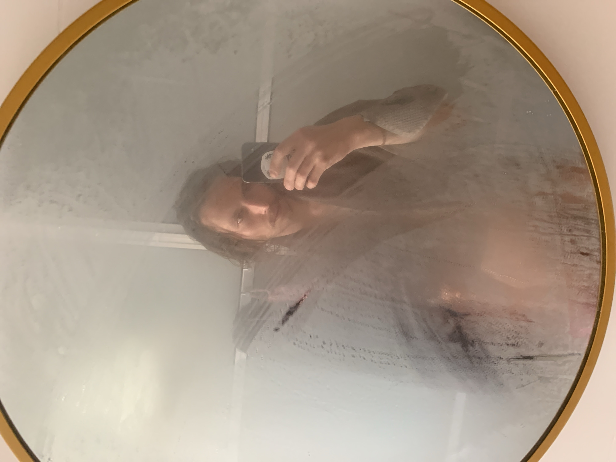 A person is taking a selfie in a steamy round mirror with a gold frame. The mirror's surface is largely covered in condensation, making the reflection obscured in places. The person is holding a smartphone in their hand, visible in the mirror. Their face, hair, and upper body are visible through the steam. A vertical line is partially wiped away from the center of the mirror. The surroundings appear to be a bathroom, with a white wall partially visible around the edge of the mirror. The location is not discernible as Tel-Aviv, Israel from the image.