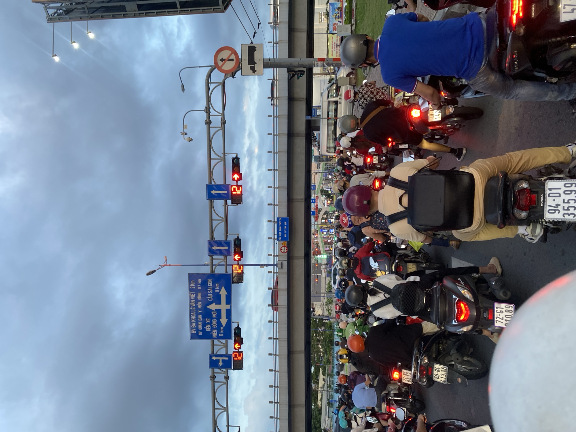 The image shows heavy traffic in Thủ Đức, Vietnam. Many people are riding motorbikes, waiting at a red light on a multi-lane road. Overhead traffic signs indicate distances to local landmarks, including BV Đa Khoa Lê Văn Việt (2 km), BV Quân Dân Y Miền Đông (0.7 km), Bến Xe Miền Đông Mới (6 km), and Cầu Sài Gòn (9 km). A sign indicates Cầu Vượt Bằng Thép Tại Ngã Tư Thủ Đức (steel overpass at Thủ Đức intersection). The sky is overcast.