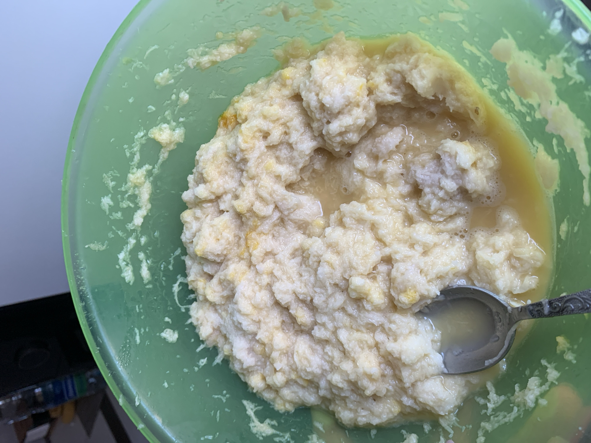 A close-up, overhead shot shows a bright green plastic bowl filled with a lumpy, off-white mixture. The mixture appears to be a food preparation, possibly a dough or batter, with visible streaks of yellow, suggesting the presence of egg yolks or other ingredients. Some liquid has pooled at the bottom of the bowl. A tarnished silver spoon rests in the mixture, its bowl partially filled with the liquid. The interior of the bowl has a slightly greasy sheen, with some of the mixture adhering to the sides. In the background, out of focus, a dark object, possibly a speaker, and a plastic bottle with a blue label are visible. The lighting is moderate, casting soft shadows. The scene is indoors, and based on the provided context, it is located in Kapyĺ, Belarus.