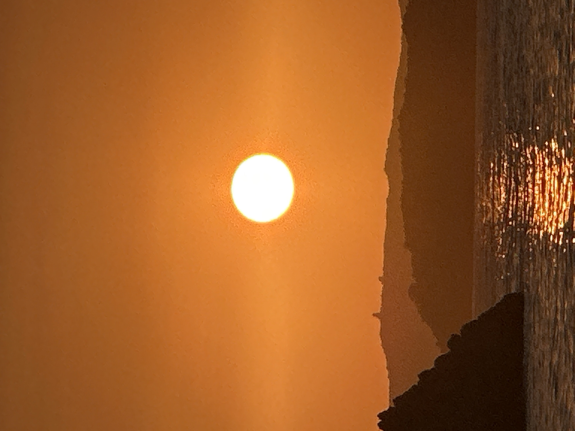This is a vertical, eye-level, close-up shot of a sunset over the Adriatic Sea, near Sveti Stefan, Montenegro. The sun, a bright white orb with a slight halo, is positioned slightly left of center in the upper half of the frame, casting a warm, orange glow across the entire scene. The sky is a gradient of deep orange at the top, transitioning to a lighter, yellowish-orange near the horizon.
In the mid-ground, dark, silhouetted hills form a layered backdrop. The closest hill on the left is a jagged, rocky outcropping. Further back, the silhouettes of other hills are visible, with a distinct, tower-like structure faintly discernible atop one of the distant peaks, likely the Fortress of Sveti Stefan.
The foreground is dominated by the calm, rippling surface of the sea. Sunlight reflects off the water in a shimmering path, creating streaks of orange and gold. The overall atmosphere is tranquil and serene, capturing the beauty of a coastal sunset.