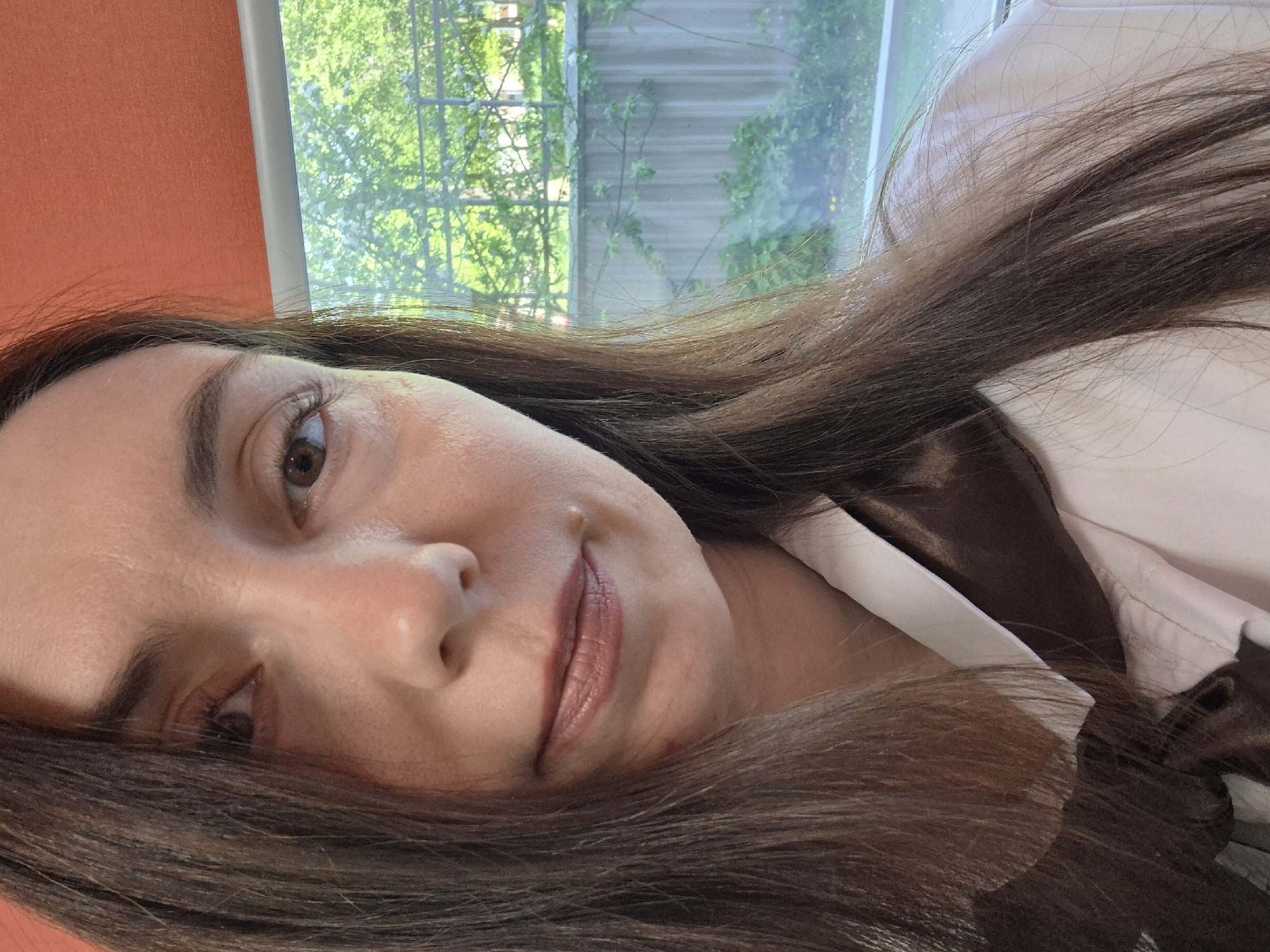 A close-up shot of a woman's face. She has brown hair, brown eyes, and a mole on her right cheek. She is wearing a white and brown top. The background includes a window with a view of trees and an orange wall. The location is Unknown, Unknown.