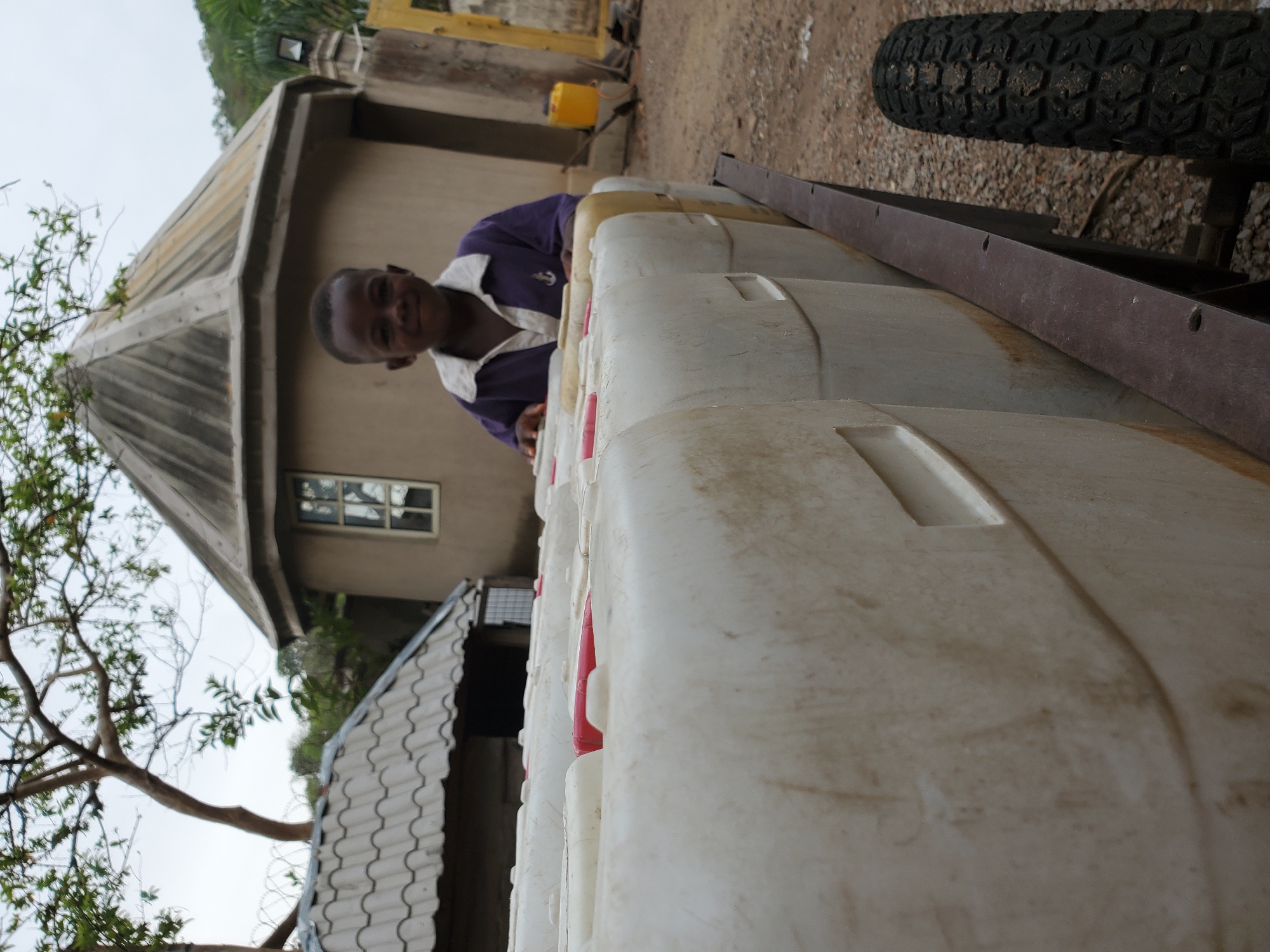 The image, taken in Mararaba, Nigeria, depicts a young person smiling behind a row of large plastic containers in an outdoor setting, with the photo rotated 90 degrees clockwise.
In the upper center of the frame, a young person, likely a boy, is seen smiling warmly at the camera. He has short dark hair and is wearing a dark purple collared shirt with white accents on the collar and a white emblem, possibly an anchor, on his chest.
The immediate foreground is filled with several large, light-colored (off-white or cream) plastic containers, some with red tops or caps, arranged in a row. To the left, a building with a distinctive conical or pyramidal roof made of corrugated material is prominent, featuring a single rectangular window. Another structure is partially visible further back on the right. A robust black tire with a textured tread and a long, dark metal bar or frame are visible on the far right side of the image. A small, sparse tree with branches extends into the frame from the far left. The ground is a mix of dirt and gravel, and a yellow container is discernible in the background behind the main row of containers. The sky appears overcast, suggesting it is daytime with diffused lighting. The young person's smile conveys a friendly and positive emotion. There is no visible text in the image.