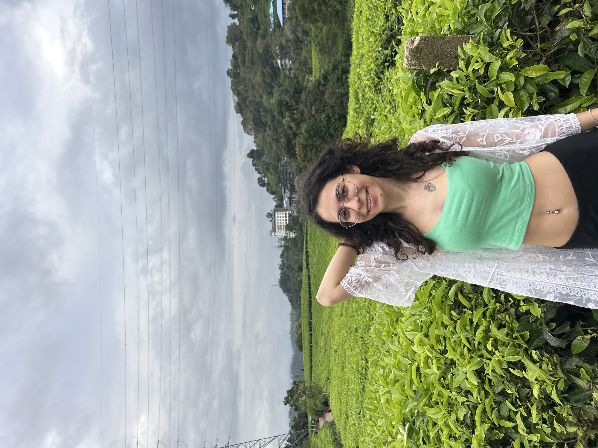 A young woman with long, dark, curly hair, round glasses, a light green crop top, black pants, and a white, lacy, sheer long-sleeved cover-up smiles broadly at the camera in this outdoor portrait. She has a small anatomical heart tattoo on her left upper chest and a belly button piercing. Her left arm is bent with her hand near her head, while her right arm is also bent.
The setting is a lush, green tea plantation, with neatly trimmed tea bushes surrounding her and extending into the background. Beyond the plantation, rolling hills covered in dense green foliage are visible. In the mid-ground, several buildings, including a prominent white multi-story structure, are nestled among the trees. The sky is overcast and grey with clouds, suggesting a cloudy day. Multiple power lines stretch across the upper portion of the frame, and a power pylon is partially visible in the lower left. The scene evokes a tranquil, natural environment, likely in a hilly region known for tea cultivation, such as Pallivasal, India. The woman appears happy and relaxed, enjoying her surroundings. There is no visible text.