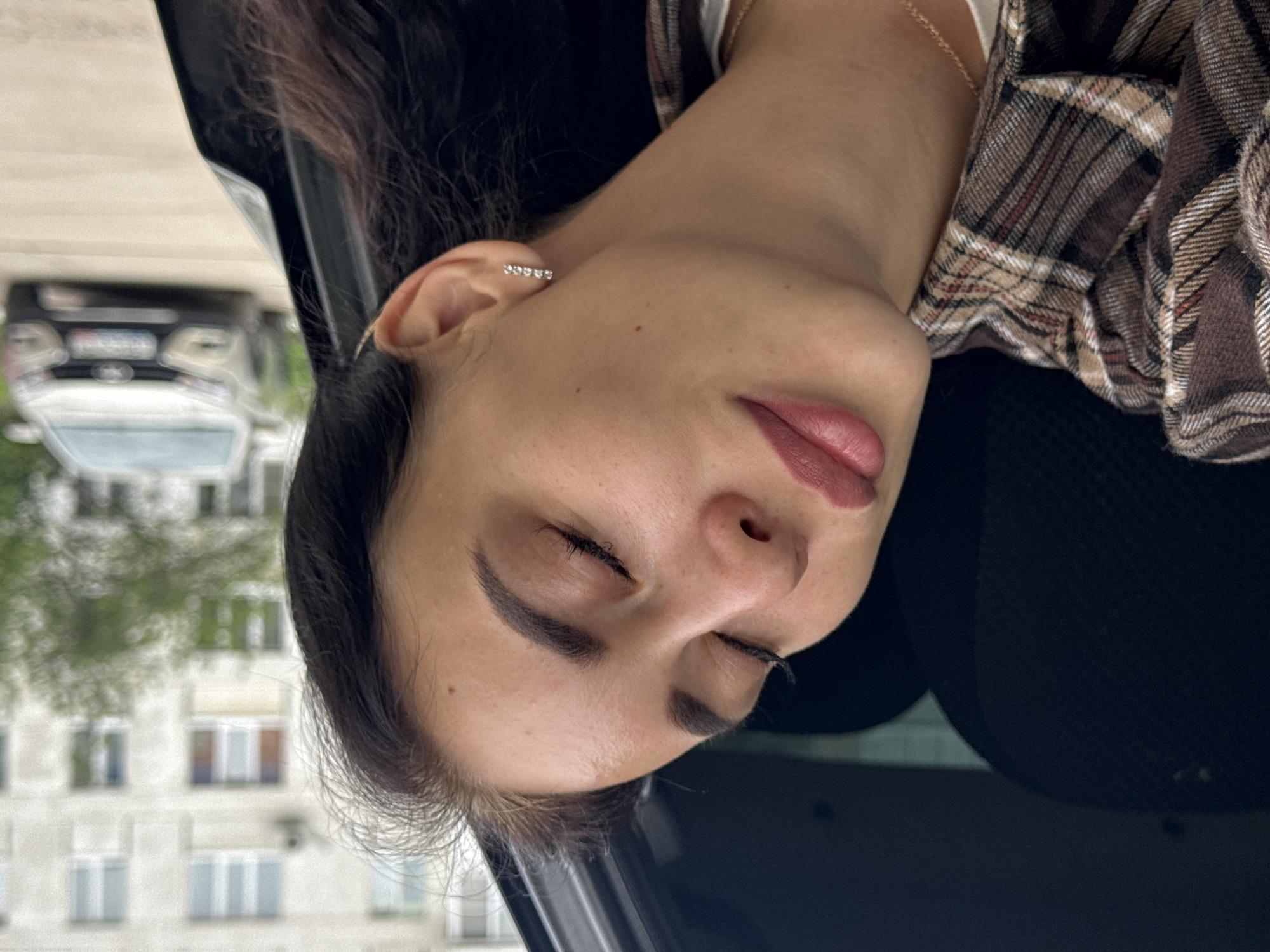 The image features a young woman resting inside a car, viewed from a close-up, slightly elevated angle. She has dark hair, styled to the side, and her eyes are closed, suggesting she is asleep or resting peacefully. Her lips are slightly parted, and she wears makeup, including filled eyebrows and dark pink lipstick. A small, linear earring with multiple shimmering stones adorns her right ear. She is dressed in a brown and beige plaid shirt over a white garment, with a delicate gold chain necklace visible around her neck.
The setting appears to be a car interior, with dark upholstery visible around her. Through the car window, an urban or suburban environment is blurred in the background. A dark-colored vehicle is partially visible and out of focus. Behind it, a multi-story building with numerous windows and a muted, light-colored facade can be seen, partially obscured by some green foliage. The lighting suggests it is daytime, possibly overcast, given the soft, diffused light and lack of harsh shadows. The overall impression is one of calm and tranquility as the woman rests while possibly commuting or waiting. There is no visible text.