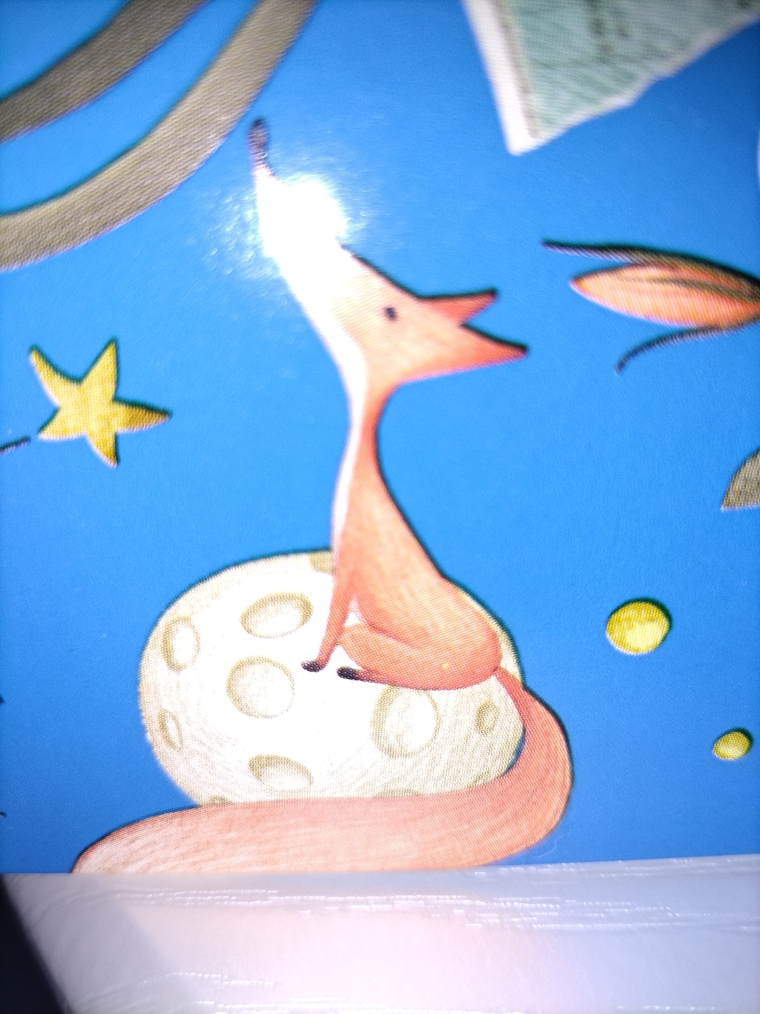 An illustration on a blue background features an orange fox sitting on a round, cratered object resembling the moon. The fox is facing left, with its head tilted upwards and its mouth open as if howling. A bright white light emanates from its snout. To the left of the fox, a yellow star with five points is visible. In the upper left, there are two curved, gray, textured bands. To the right of the fox, there are two small yellow circles, and a pointed, orange and black object with a curved tail. The bottom of the image shows a white, textured surface.