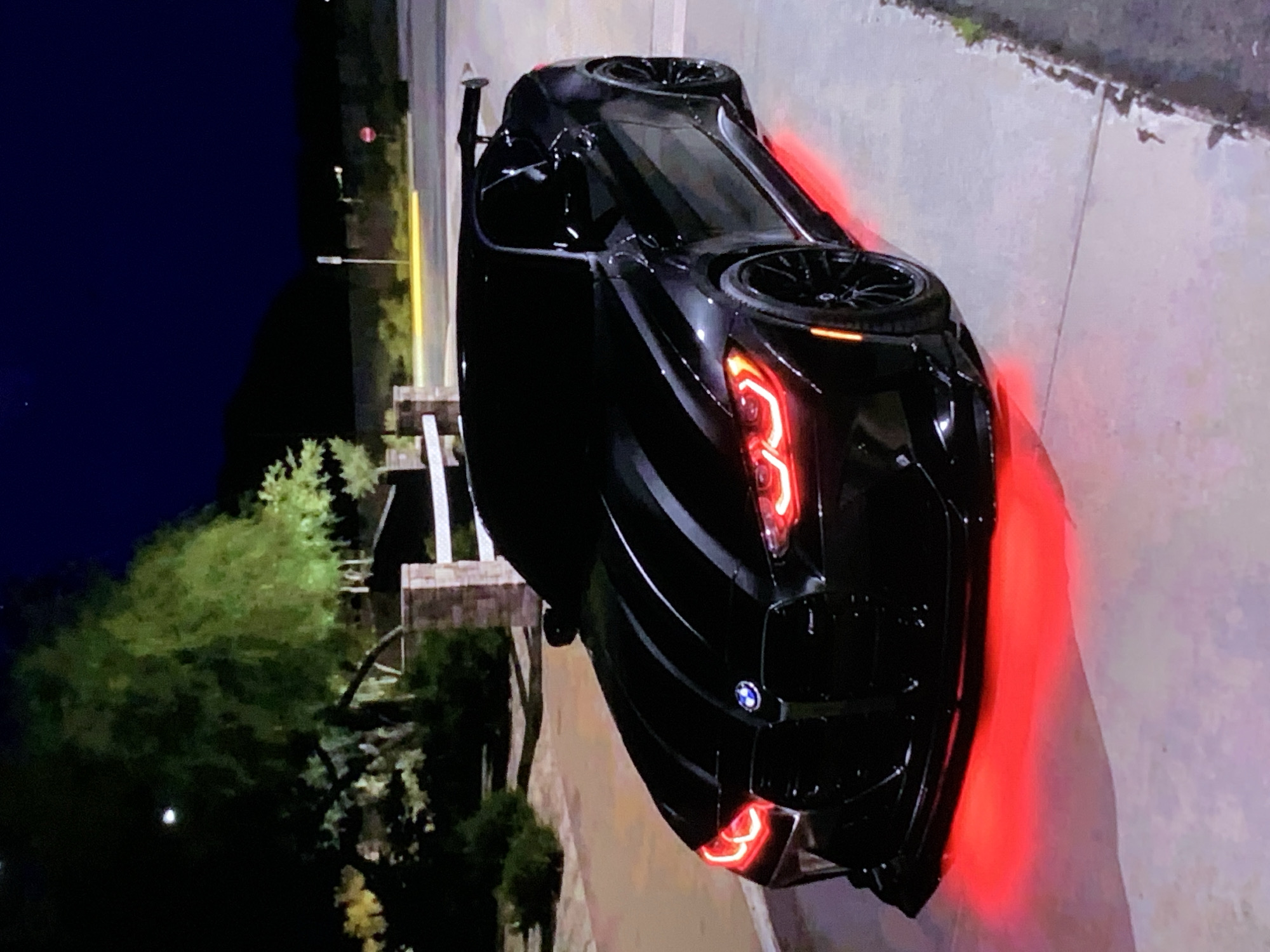 A black BMW M4 Coupe is parked at night on a paved surface with red underglow lighting. The car has a large rear wing and red accents on its headlights. In the background, there is a stone pillar, a white fence, trees, and a dark mountainous landscape under a night sky. The scene appears to be in a residential or suburban area.