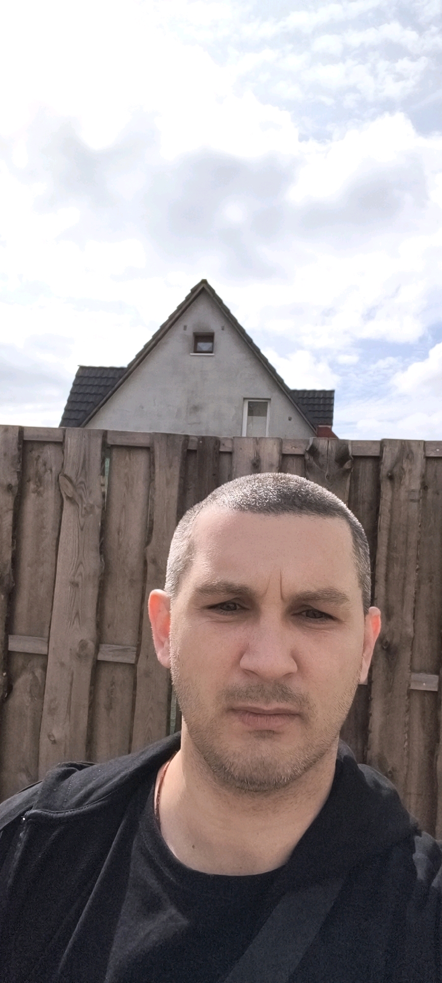 A man in a black jacket stands in front of a wooden fence, with a house visible in the background under a cloudy sky. The man's head and shoulders are in the foreground, and he is looking at the camera. The house is a typical German residential building with a gable roof and a small window. The fence is made of weathered wooden planks. The scene appears to be in an outdoor residential area.