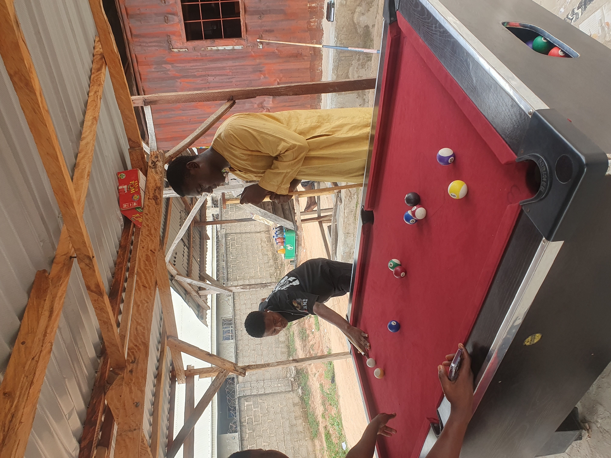 The image depicts an outdoor pool table area in Lokoja, Nigeria. The scene is under a makeshift roof constructed of corrugated metal sheeting supported by wooden beams. Three men are present. One man, wearing a black t-shirt with a graphic print, is actively playing pool, leaning over to strike a white cue ball with a cue stick. Another man, dressed in a yellow kaftan, stands a short distance away, holding a cue stick and observing the game. The third person's arm is visible on the left side of the frame, reaching towards the pool table. The red felt surface of the pool table is scattered with several billiard balls of various colors and numbers. In the background, a light grey brick wall with barbed wire runs along the perimeter, suggesting a secure or private area. Beyond the wall, a landscape with sparse vegetation is visible. A red corrugated metal structure with a barred window stands to the right of the pool table. The overall environment suggests a casual recreational setting.