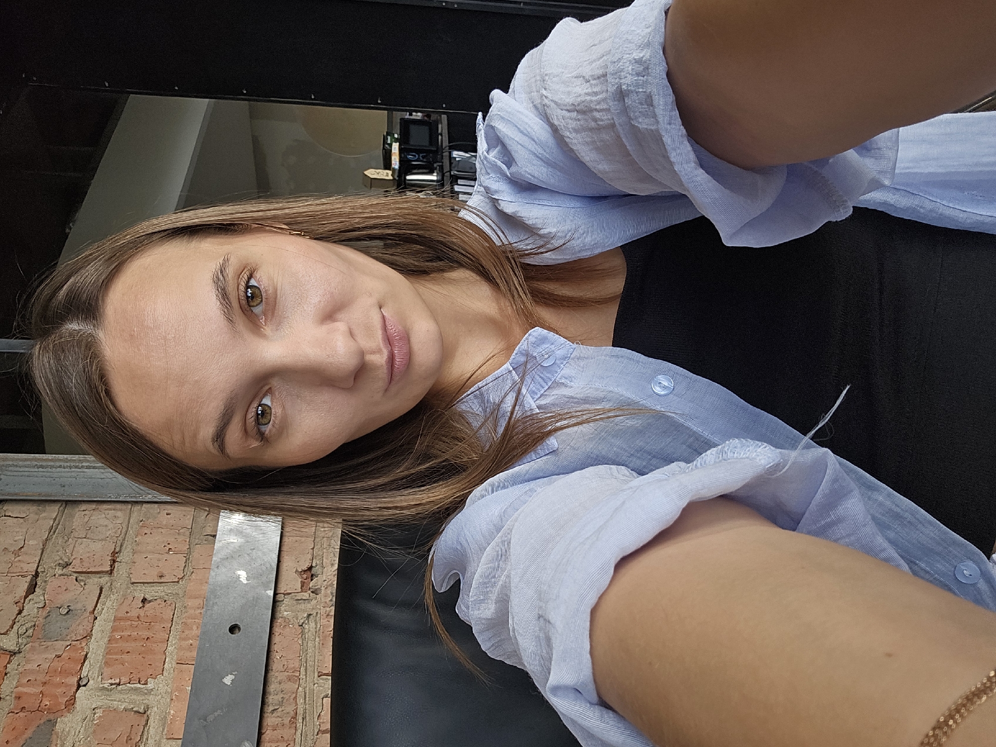 A young woman with brown hair is taking a selfie indoors, seated in a dark chair in front of a brick wall. She is wearing a light blue button-down shirt over a black top, with a gold chain visible around her neck. Her expression is neutral, with her gaze directed at the camera. The background shows a brick wall, a metal beam, and shelves with various items. The scene appears to be in an apartment or a cafe.