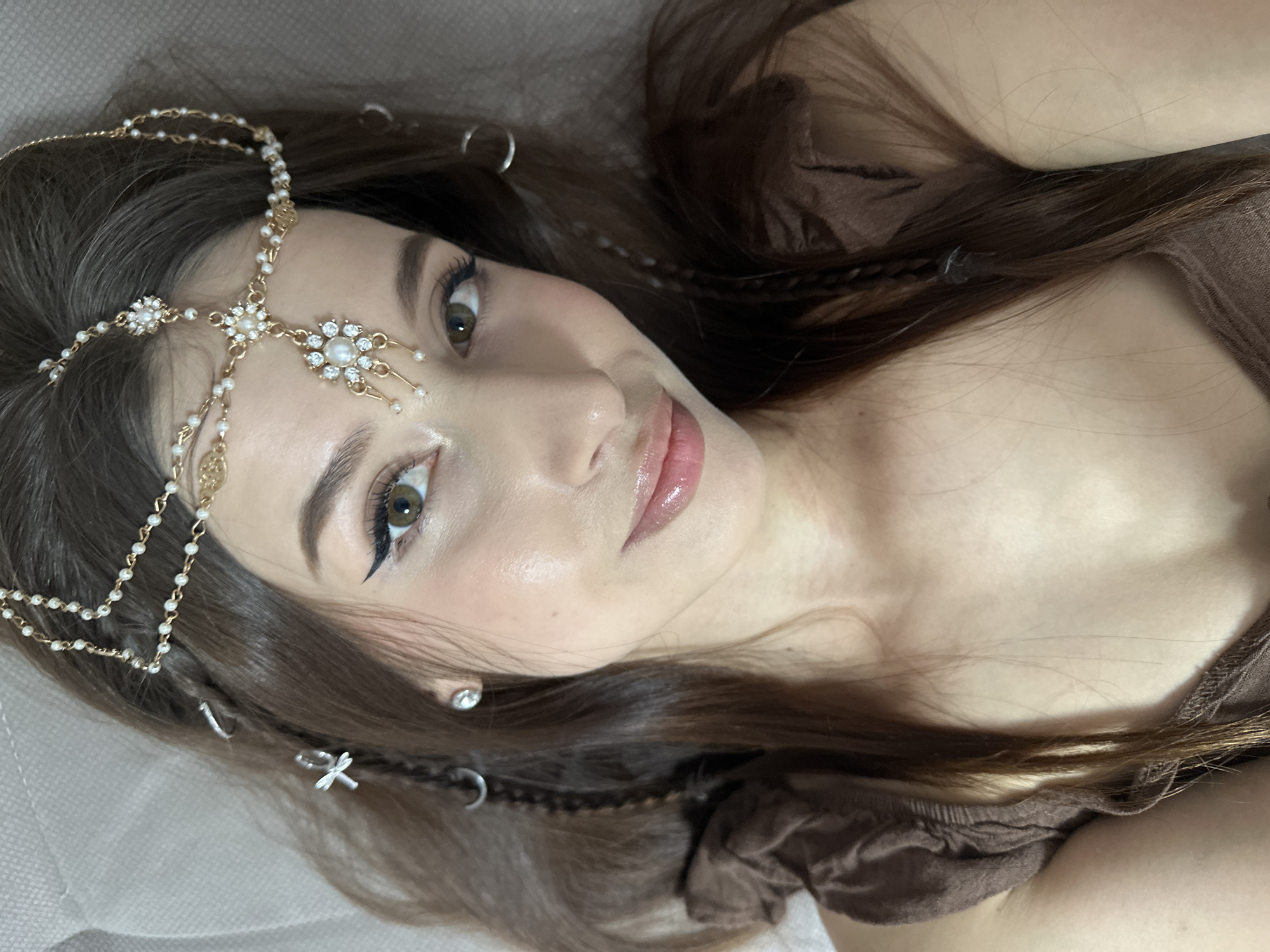 A young woman with long, flowing dark brown hair is captured in a close-up, eye-level portrait, lying down and looking towards the camera with a gentle gaze. Her head is angled slightly to her left, resting on a light-colored, soft surface.
She is wearing an elaborate gold-toned headpiece adorned with small pearls and clear rhinestones. This accessory features a central floral-like element on her forehead, with chains extending back into her hair and decorative drops resting near her temple. Her hair is further accessorized with small, silver-toned charms, including crescent moon shapes and a cross or dragonfly-like design, interspersed among thin braids. A simple clear stud earring is visible in her left ear.
Her makeup includes prominent winged eyeliner, well-defined eyebrows, and glossy, light pink lips. Her skin appears smooth and radiant. She is wearing a simple brown garment, possibly a top or dress, visible around her shoulders and chest.
The activity depicted is a posed photograph, with the woman conveying a serene and calm emotion. No specific time of day or weather can be discerned from the indoor setting. No specific location cues are visible within the image, though the provided context indicates Yekaterinburg, Russia. There is no visible text in the image.