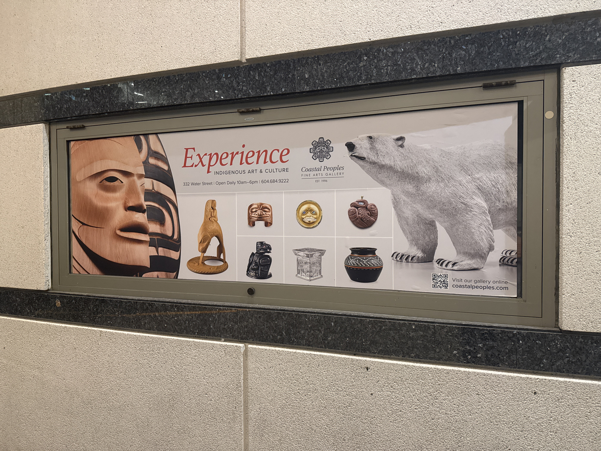 A framed advertisement for the Coastal Peoples Fine Arts Gallery is mounted on a wall. The poster contains a collage of images depicting Indigenous art and culture, including a carved wooden face, various sculptures, and a polar bear. The words "Experience Indigenous Art & Culture" are prominently displayed, along with the gallery's address at 332 Water Street in Vancouver, Canada, and its website. The wall's texture is visible around the frame, and the mounting appears to be indoors.