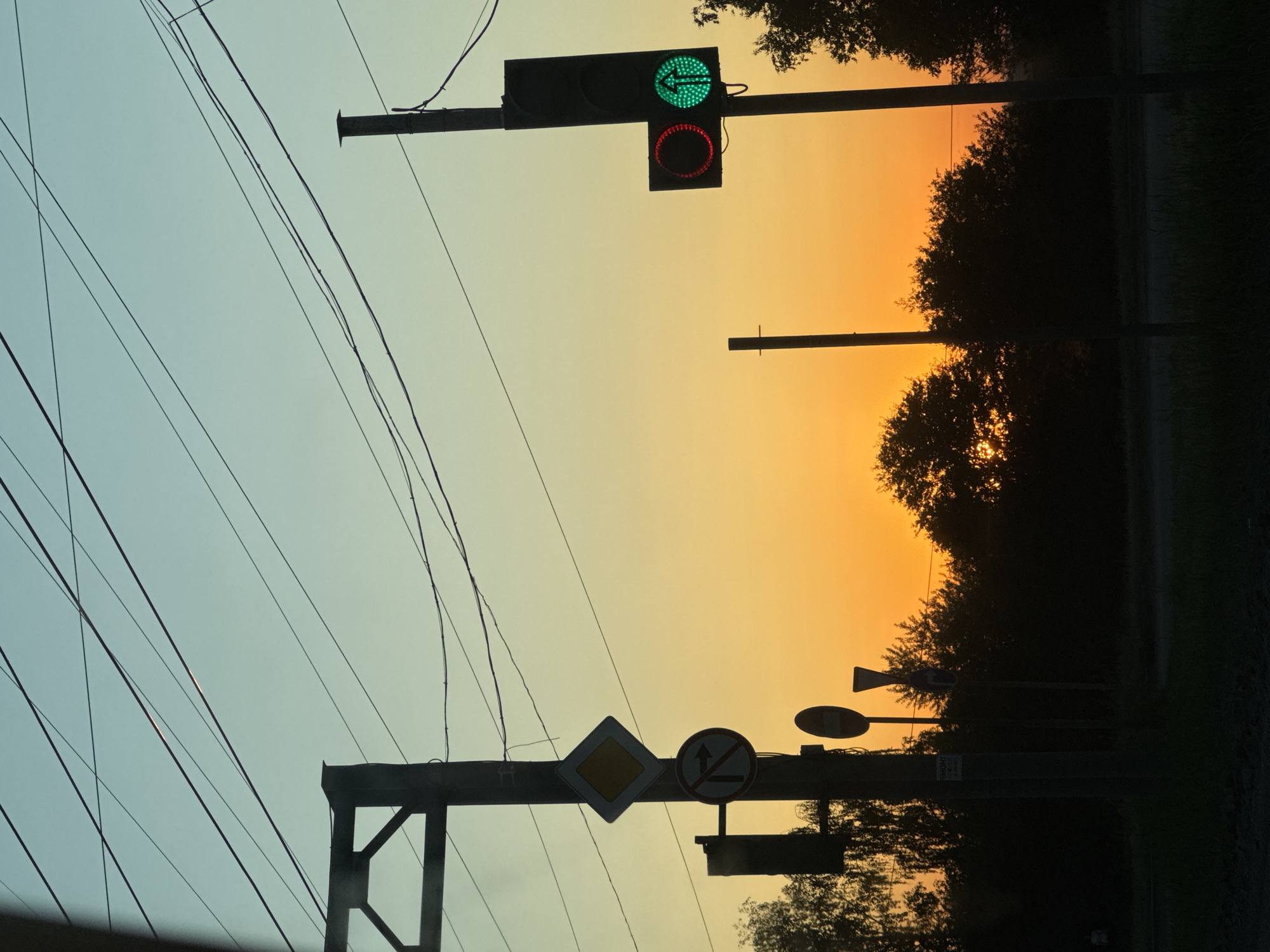 The image presents a street scene at dusk or dawn. The sky transitions from a pale blue-gray to an orange-yellow hue. A traffic light is visible with a green left-turn signal and a red light beneath it. Below, a road sign structure supports multiple signs, including a yellow diamond-shaped sign and a round sign with a horizontal line bisecting an arrow pointing upwards, possibly indicating a prohibition. Electrical wires crisscross the sky. The foreground is slightly darker, with outlines of foliage suggesting trees and bushes. In the background, darker shapes of trees and other roadside objects can be discerned against the bright sky. The location is unidentified, but it appears to be an urban or suburban street with typical road infrastructure.
