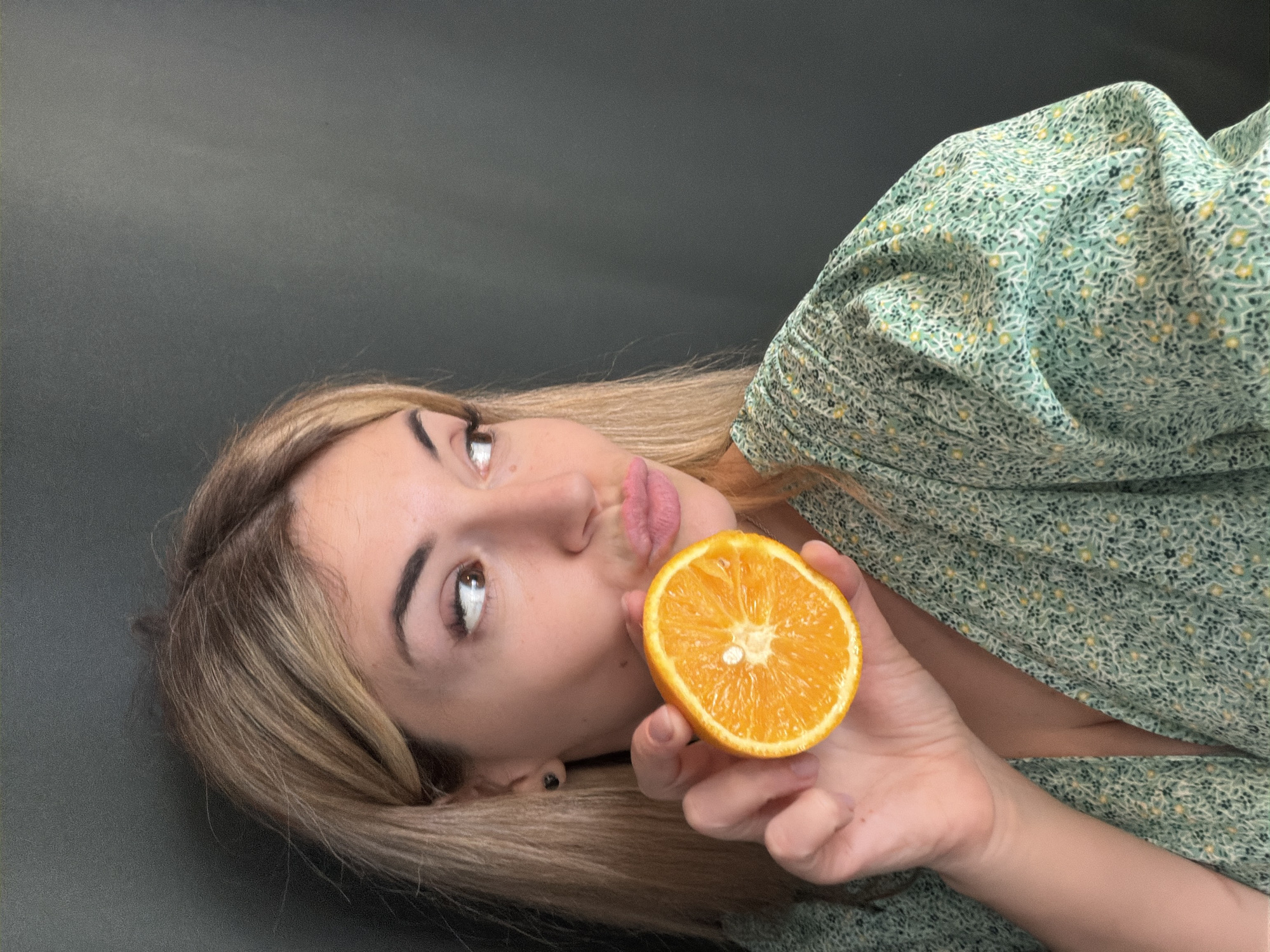 A close-up, eye-level shot captures a young woman lying down against a dark grey background, with her head tilted to the right. She has long, straight blonde hair, dark eyebrows, and light-colored eyes, which are looking upwards and to the left. Her lips are puckered into a "kissing" or "duck face" expression.
She is holding half an orange in her right hand, positioned near her mouth. The orange is cut cleanly, revealing its vibrant orange flesh, segments, and a small white seed in the center. She wears a light green dress or top with a delicate pattern of tiny yellow, white, and dark green flowers. A black earring is visible in her right ear.
The lighting is soft and even, suggesting daytime. The overall mood of the image is playful and quirky. No specific location cues are visible within the image, but the location context provided is Budva, Montenegro. There is no visible text in the image.