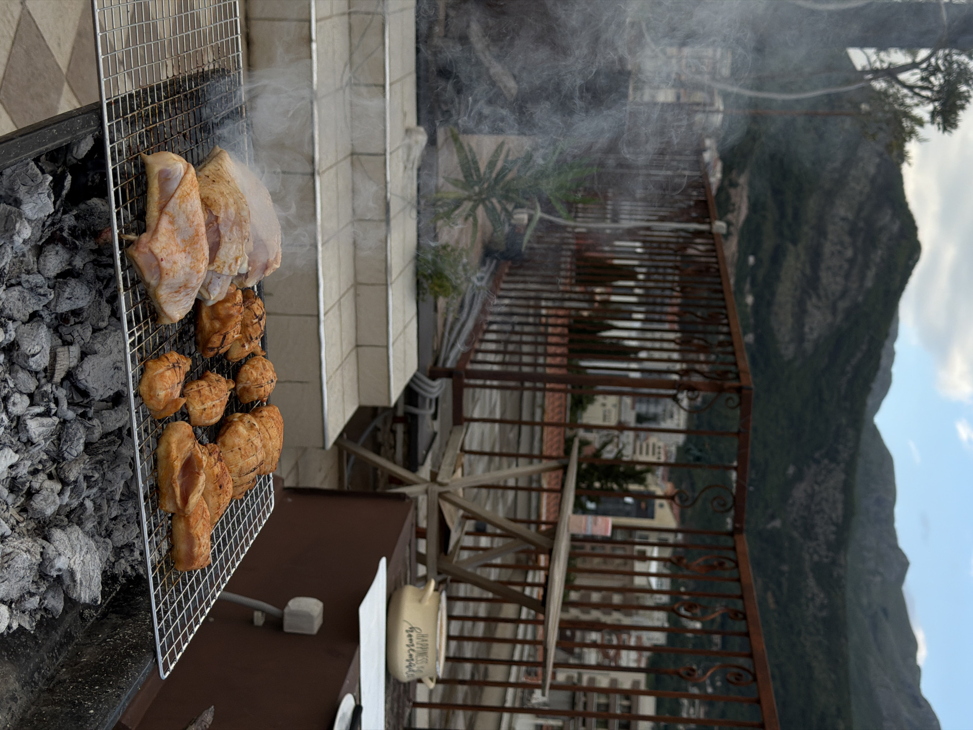 The image depicts food being grilled outdoors, with a view of the city of Budva, Montenegro and the surrounding mountainous landscape.
In the foreground, a grill containing charcoal and a metal grate holds both raw and cooked chicken. Smoke rises from the grill.
The grill is positioned on a balcony or patio, indicated by a railing and partial view of a building. Beyond the railing, the cityscape of Budva is visible, with buildings densely packed along the coastline. Behind the city, a large, green mountain dominates the background. The sky is partly cloudy.
The scene suggests a domestic setting, with someone preparing food in an outdoor space with a scenic view.