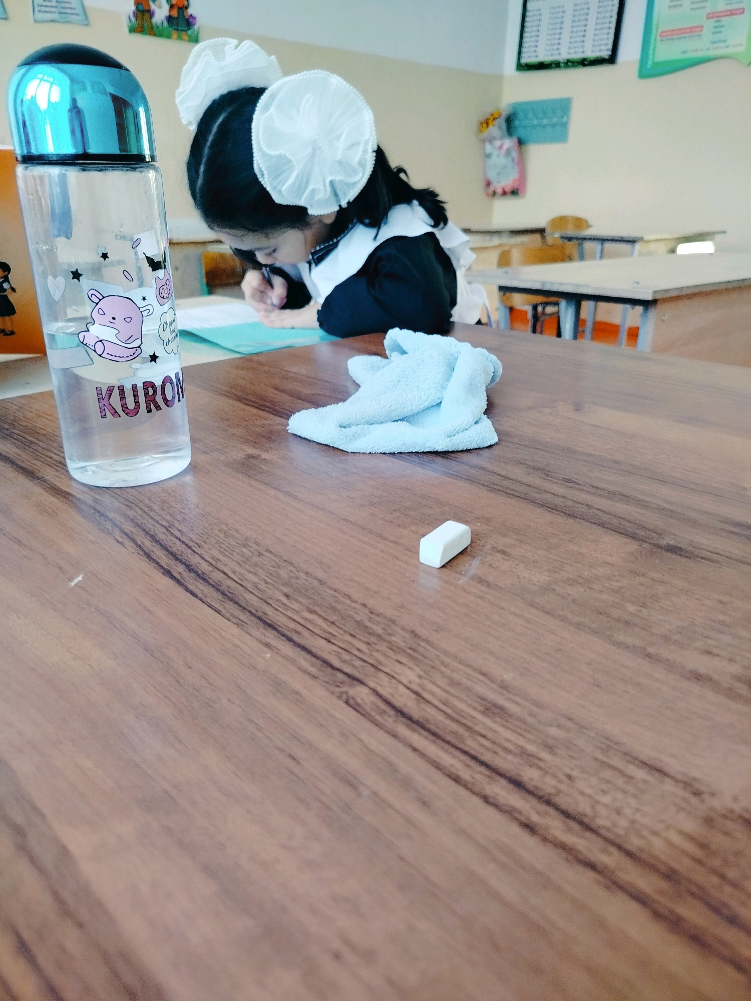 A young girl in a school uniform with large white hair bows is writing at a wooden desk. She is in a classroom, with other desks visible in the background. On the desk in front of her are a water bottle with cartoon drawings and the word "KURON" on it, a light blue folded cloth, and a small white eraser. The classroom walls are light-colored, and there are posters and educational materials on them. The lighting suggests it is daytime. The specific location is identified as a classroom in the city of Көлкент, Kazakhstan.