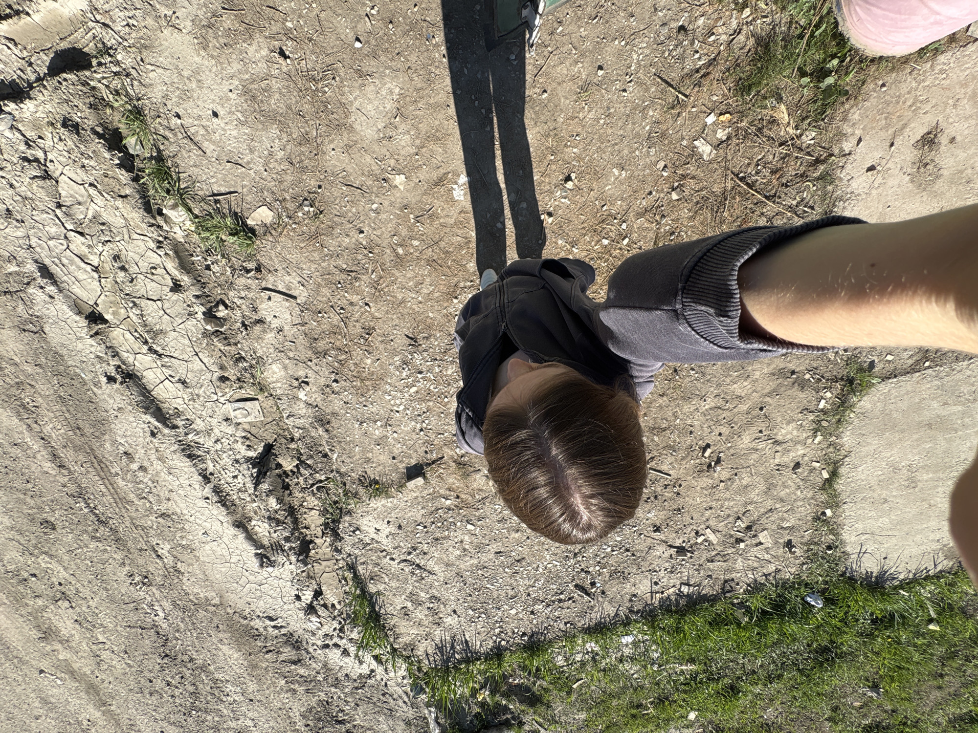 A top-down view depicts a person standing on a dirt and grass ground. The person is wearing a black jacket with grey sleeves. Part of their head and shadow are visible. A pink shoe is seen in the periphery. The ground is a mix of dry, cracked earth, loose dirt, and patches of green grass. The location is potentially in Perm, Russia, but there is no contextual information in the image to confirm this. The scene suggests an outdoor, possibly rural or undeveloped area.