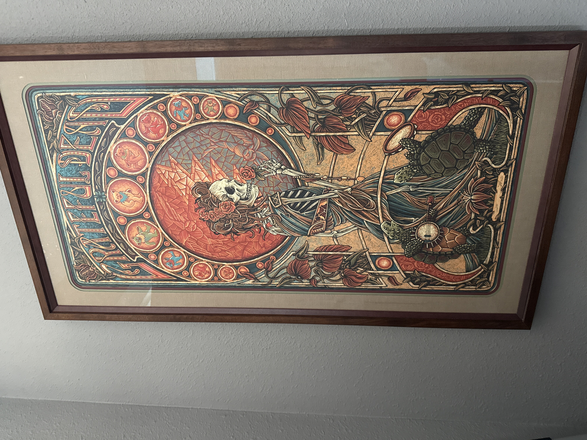 The image shows a framed poster of the Grateful Dead hanging on a textured gray wall. The poster is framed in dark wood and features a detailed illustration in an art nouveau style. The central figure is a skeleton wearing a dress and holding a rose. Surrounding the skeleton are various decorative elements, including bears, turtles, and floral motifs. The poster has a metallic sheen, suggesting it might be a foil print. The wall has a slightly rough, painted texture. The scene appears to be indoors, likely in a residential or commercial setting. There is no discernible location in the city of Tacoma, United States of America.