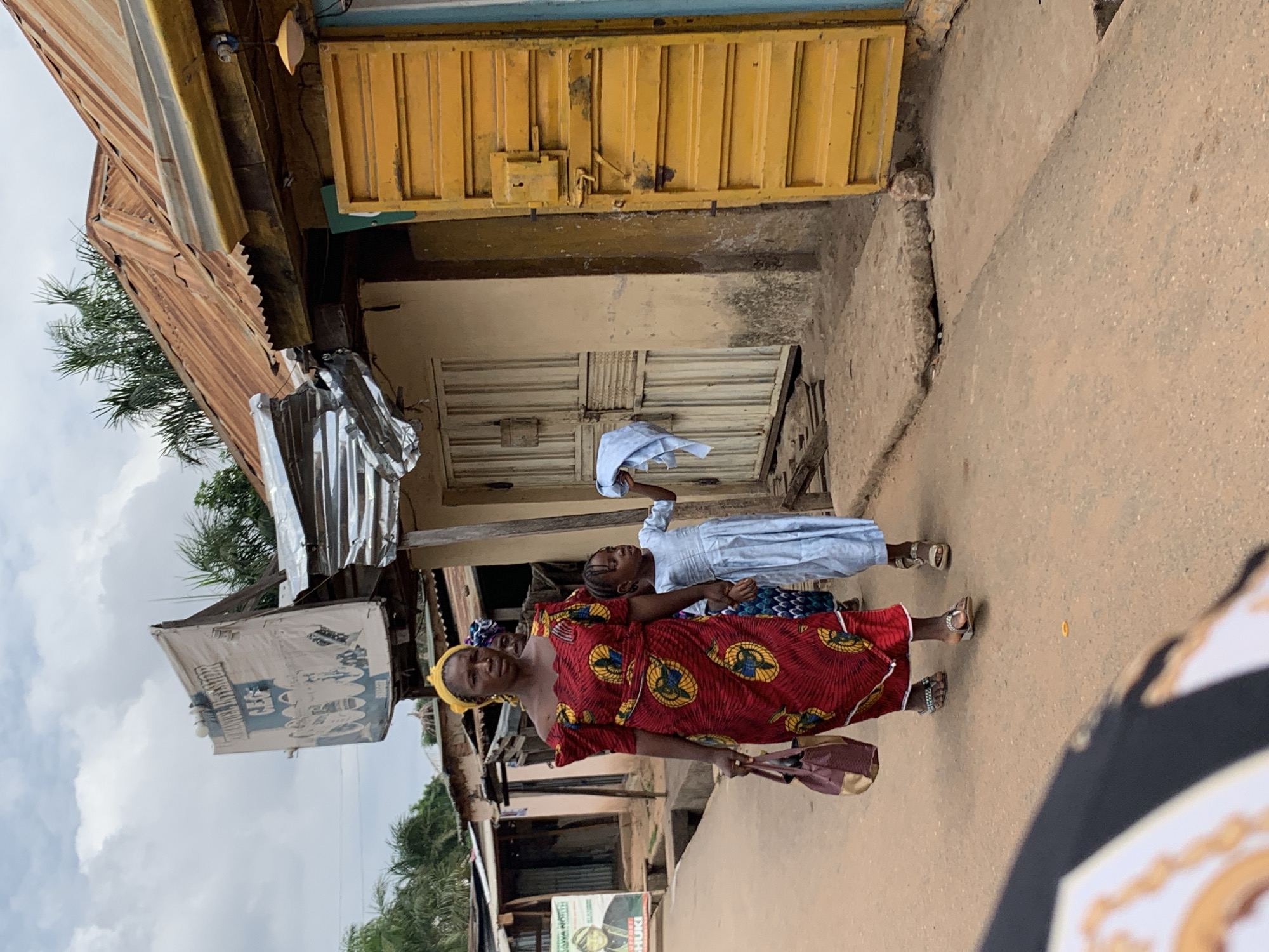 A woman and a child stand on a concrete pathway adjacent to buildings in Akwanga, Nigeria. The woman is wearing a red dress with yellow patterns, a yellow head covering, and sandals, and she is holding a purse. The child is wearing a light blue dress and sandals and is holding a white cloth in the air. The buildings are a mix of construction types, including corrugated metal, concrete, and painted wood. There is a large advertisement displayed on the side of one building.
