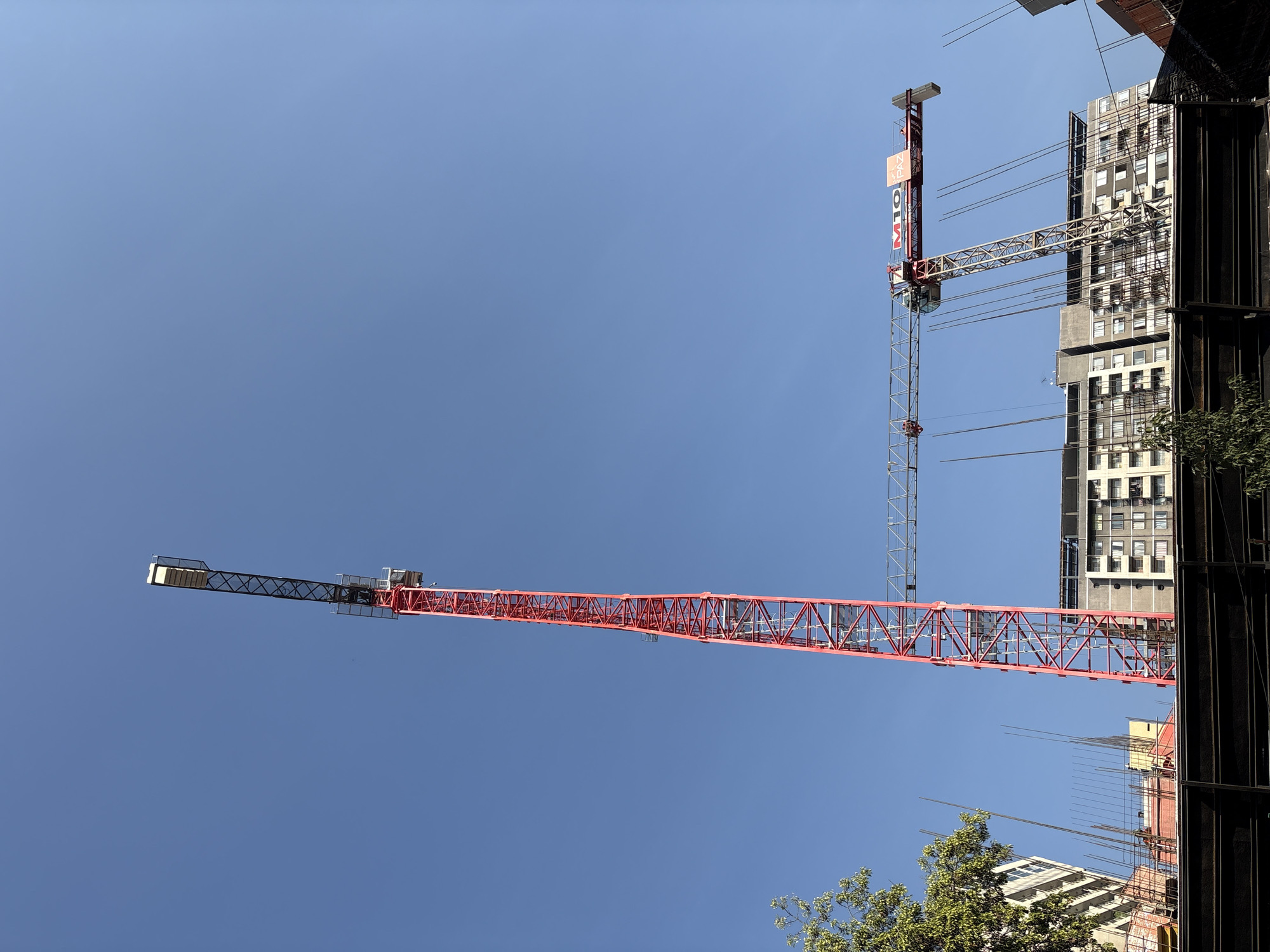 The image captures a construction site in Santiago, Chile, dominated by a tall, red tower crane. The crane's vertical arm extends high into the clear blue sky, while its horizontal jib stretches out to the right, forming an "L" shape. A second, smaller crane is attached to the jib of the larger one. Below the cranes, a multi-story building with numerous windows is visible, suggesting ongoing construction or development. The foreground shows scaffolding and construction barriers, hinting at the active nature of the site. The overall scene is one of urban development under a bright, sunny sky.