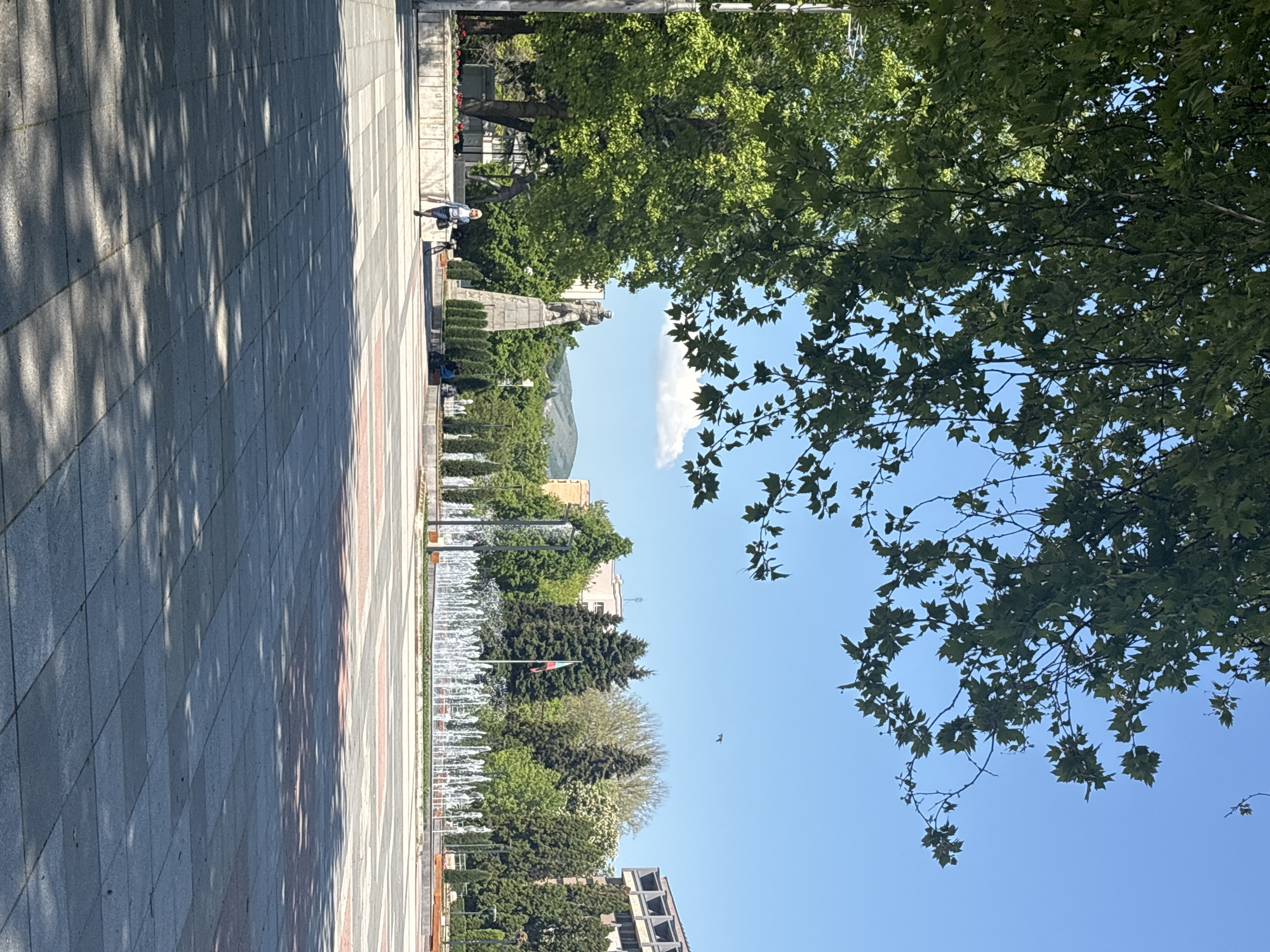 The image presents a scene in Montana, Bulgaria. The perspective is from ground level, looking along a pedestrianized area with a paved surface. The pavement is light gray and red, with a pattern that suggests individual stones or tiles. Strong shadow patterns are cast on the pavement, indicating bright sunlight.
To the right of the walkway, there's a row of trees, their foliage green and dense, partially obscuring the sky. Through gaps in the trees, a clear blue sky is visible. Further down the walkway, fountains are visible, suggesting a park or plaza area. A Bulgarian flag is visible, positioned amidst the trees.
In the distance, a monument or statue stands prominently, with what appears to be a mountain visible behind it. A single person is seen walking near the statue, appearing to be an older man wearing a hat. Modern buildings are visible along the horizon.