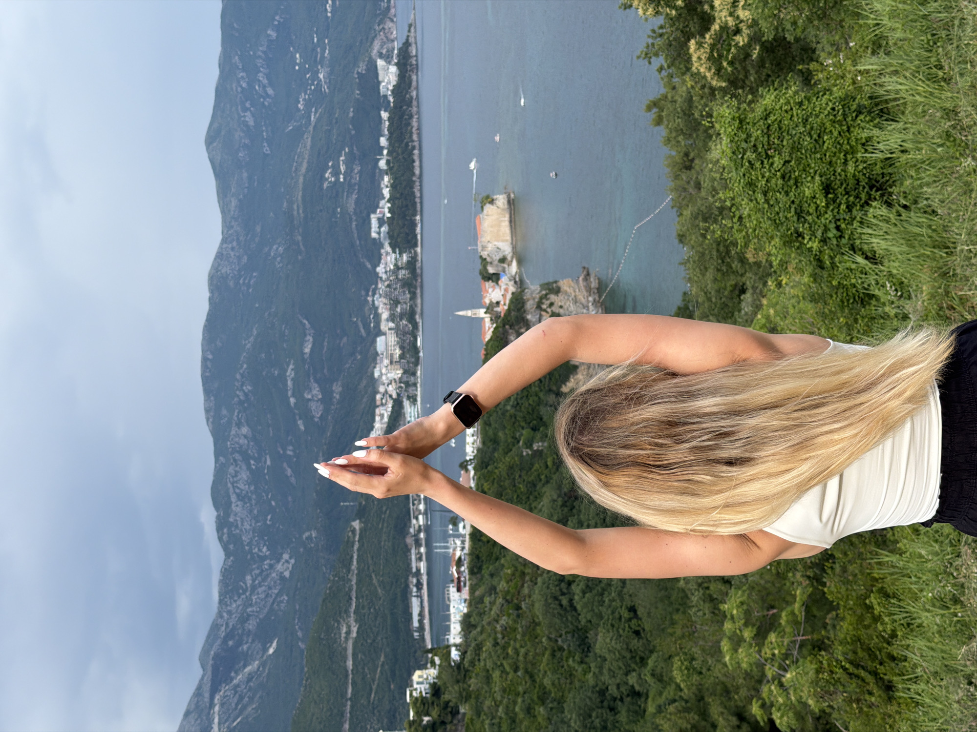 A medium-shot, vertical photo showcases a woman with long, blonde hair standing atop a hill overlooking a mountainous coastline and cityscape. Her arms are raised and outstretched. She wears a white tank top and black pants, with a black smartwatch visible on her left wrist. The city of Budva, Montenegro, is visible in the background, nestled between the mountains and the sea. The water is calm with boats docked. Lush greenery covers the hilltop and the surrounding landscape. The sky above the mountains is slightly overcast.