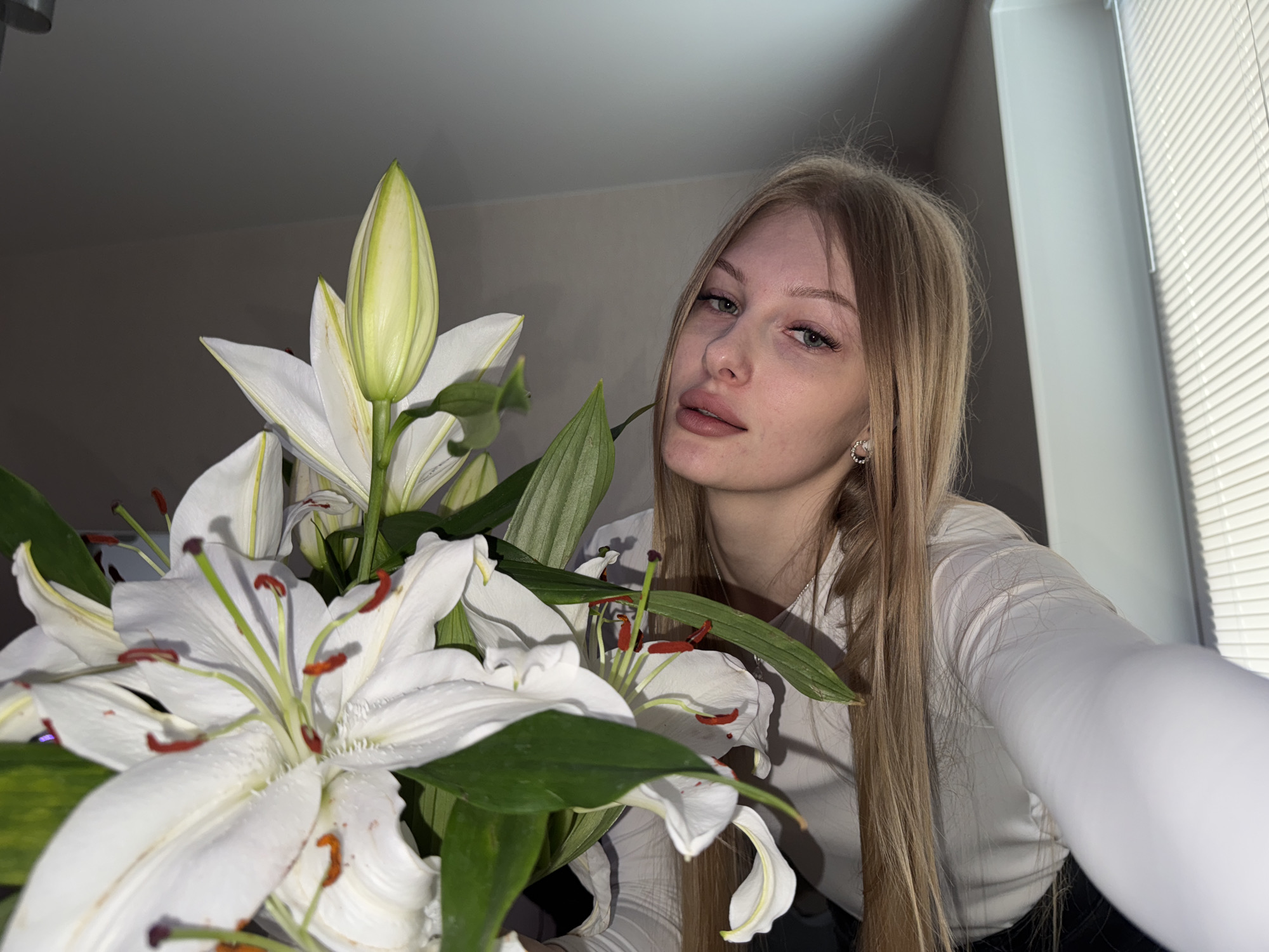 A young, fair-skinned woman with long, blonde hair stands indoors, possibly in a residential setting in Perm, Russia. She is wearing a white long-sleeved shirt and posing with a bouquet of white lilies in the foreground. The flowers have pronounced stamens with red-brown tips. The background includes a light-colored wall and a window with closed blinds. The lighting appears to be artificial.