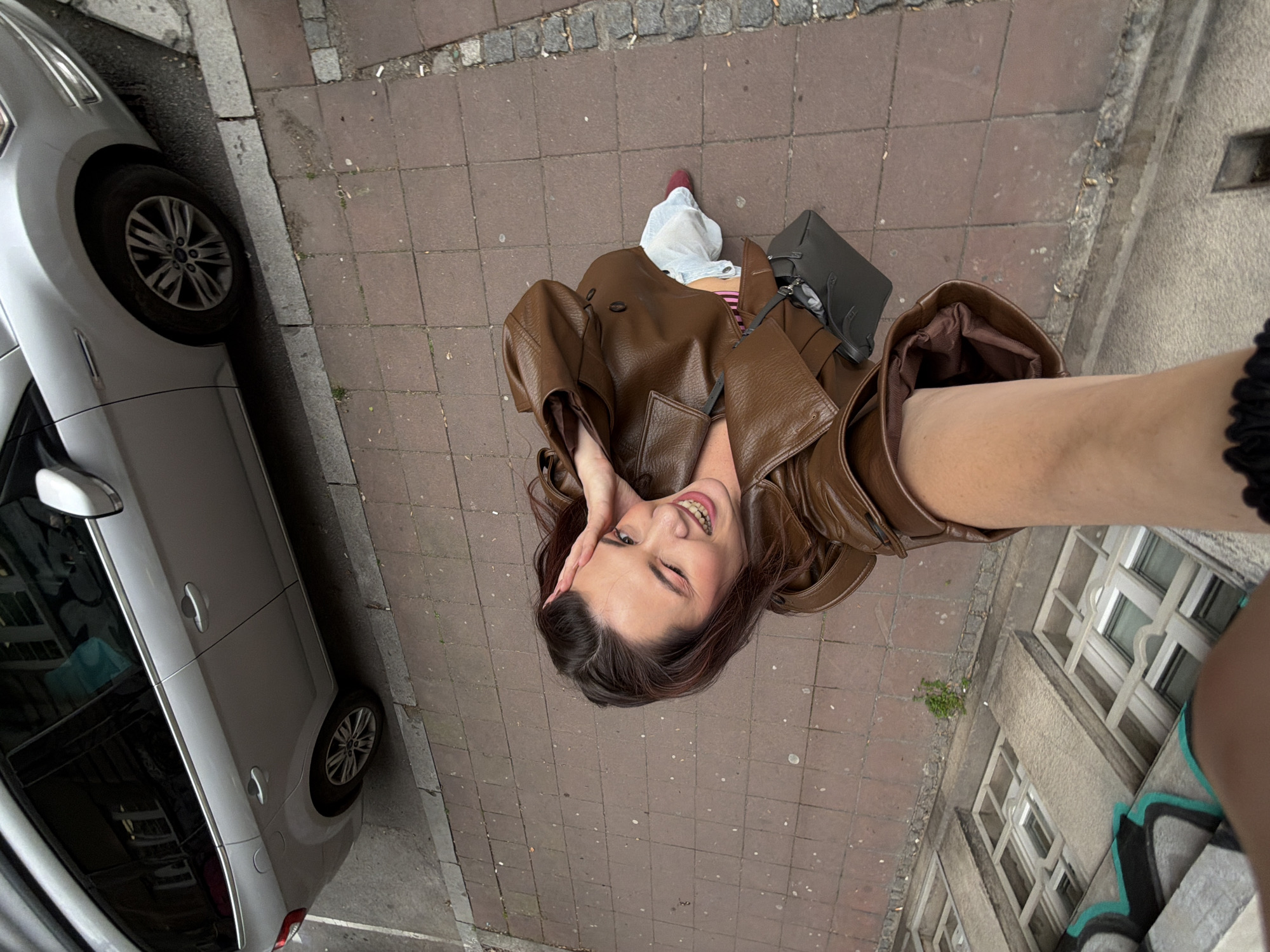 A woman in a brown leather jacket takes a selfie on a sidewalk in Belgrade, Serbia. A gray car is parked on the left side of the frame, partially obscuring the view of the street. The sidewalk is paved with square reddish-brown stones. Behind the woman, a gray building with white-framed windows is visible. Graffiti decorates the lower section of the building's wall. The woman is smiling and winking as she holds the phone to take the photo. A gray purse is also visible on the ground next to the woman.